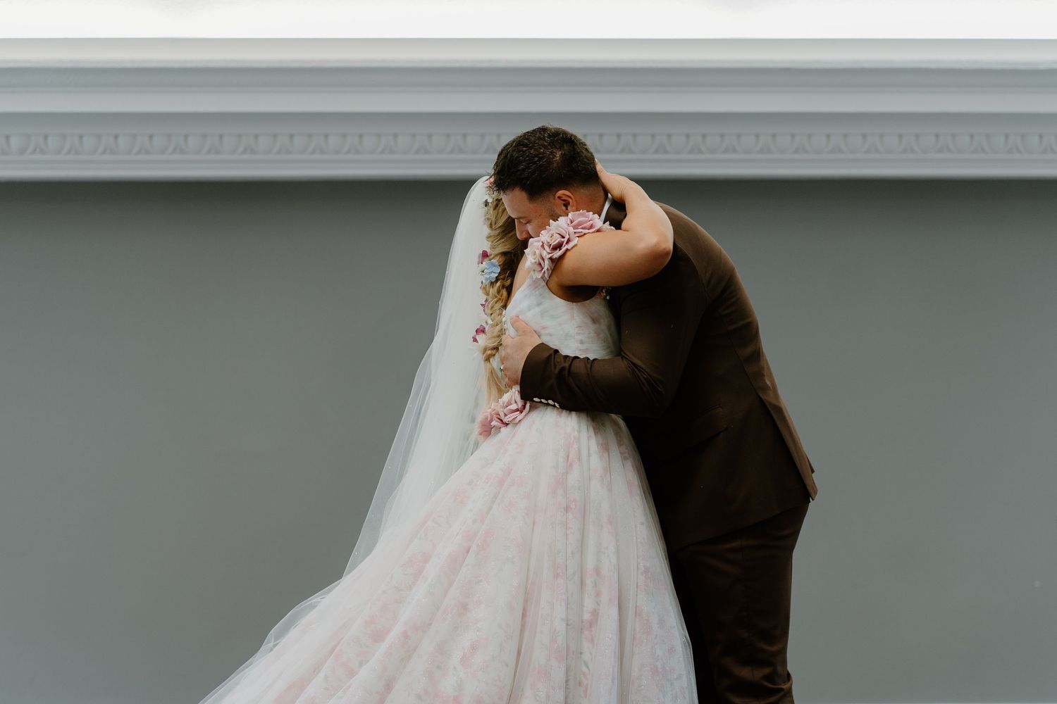 Bride and groom share a romantic embrace, her floral veil cascading down her blush wedding gown.