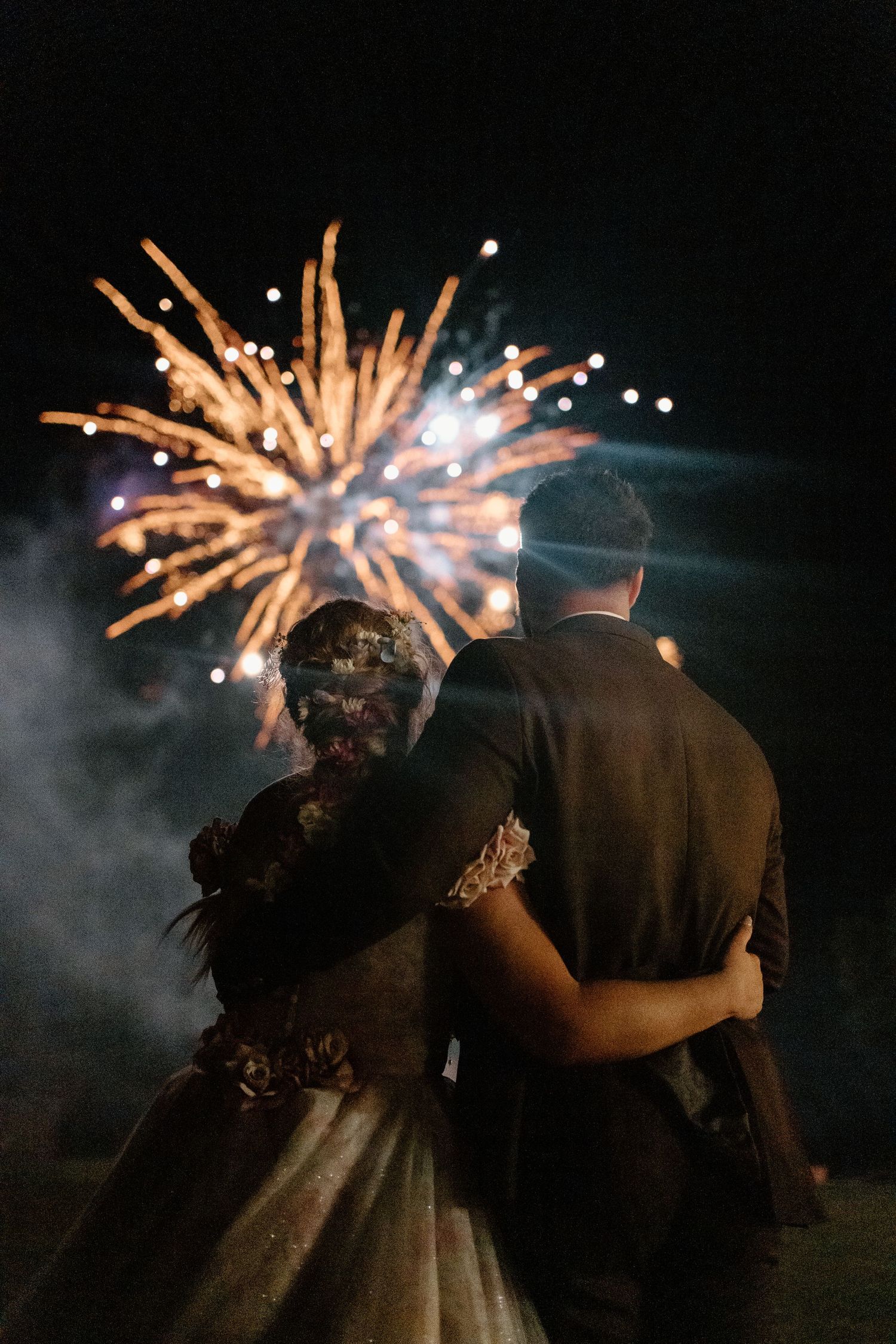 Bride and groom embracing from behind as they watch golden fireworks explode at their wedding.