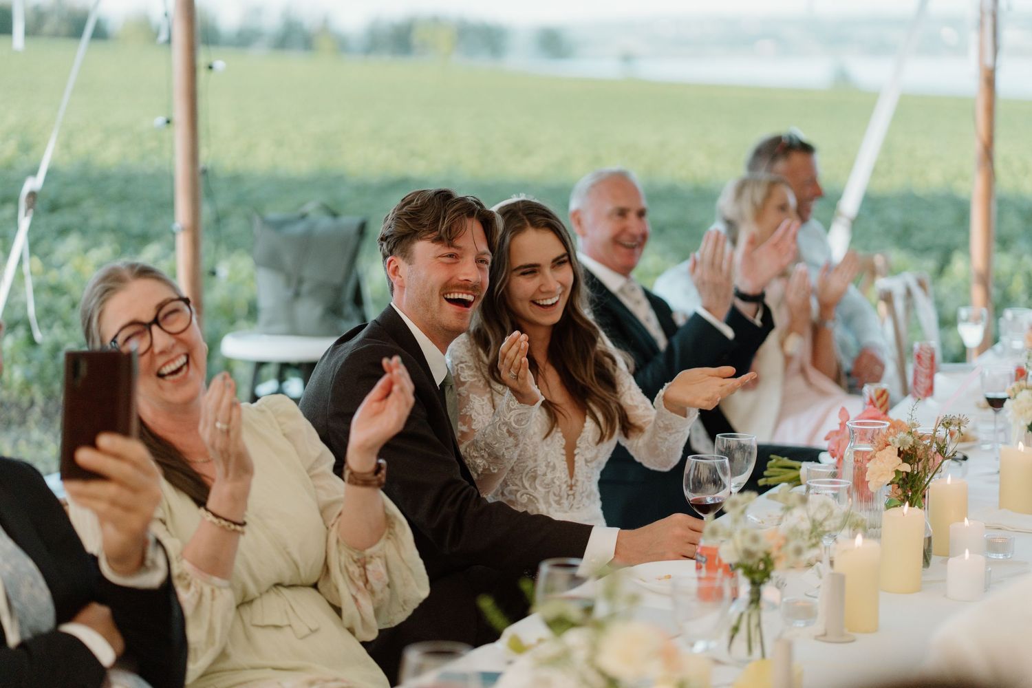 Bride and groom clapping and smiling with guests during wedding reception dinner.