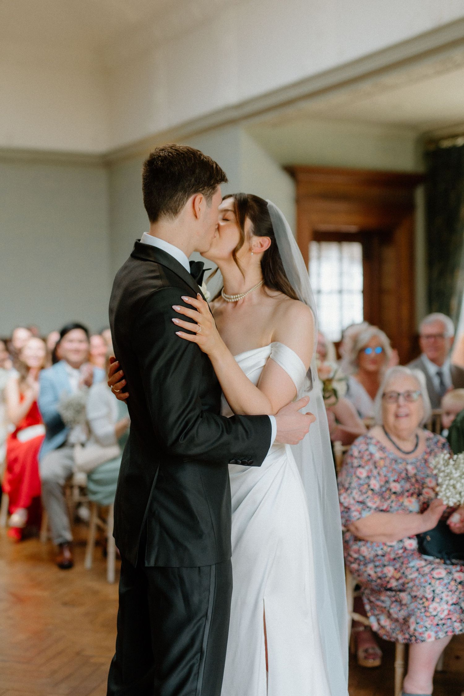 Bride and groom share first kiss at wedding ceremony as seated guests applaud in background.