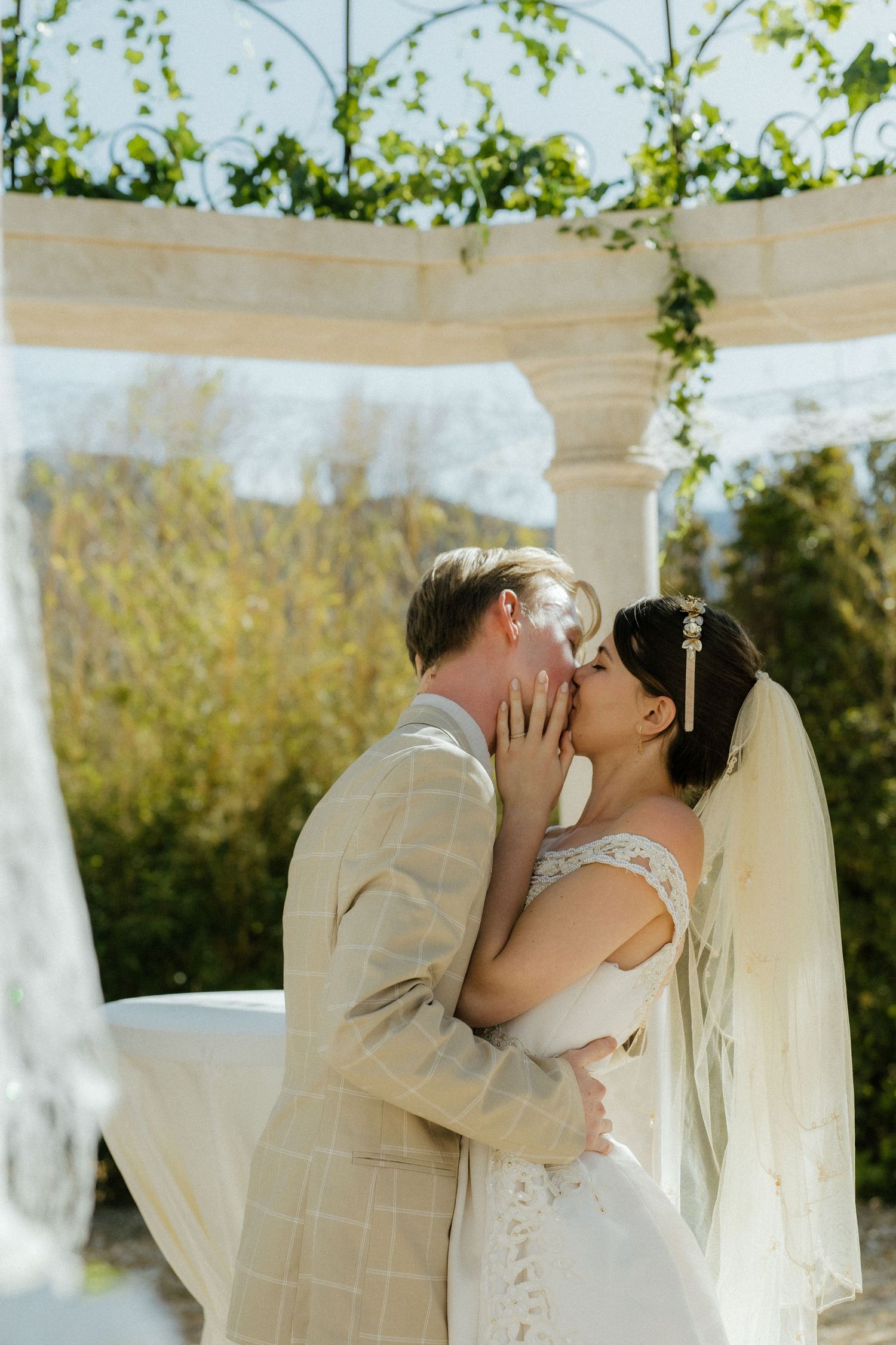 Bride and groom sharing first kiss under floral wedding arch decorated with greenery outdoors.