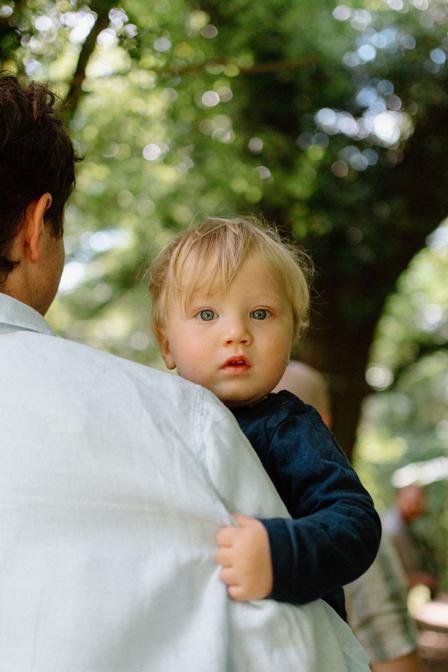 A toddler with blue eyes peeks over a parent's shoulder outdoors in a lush green park.