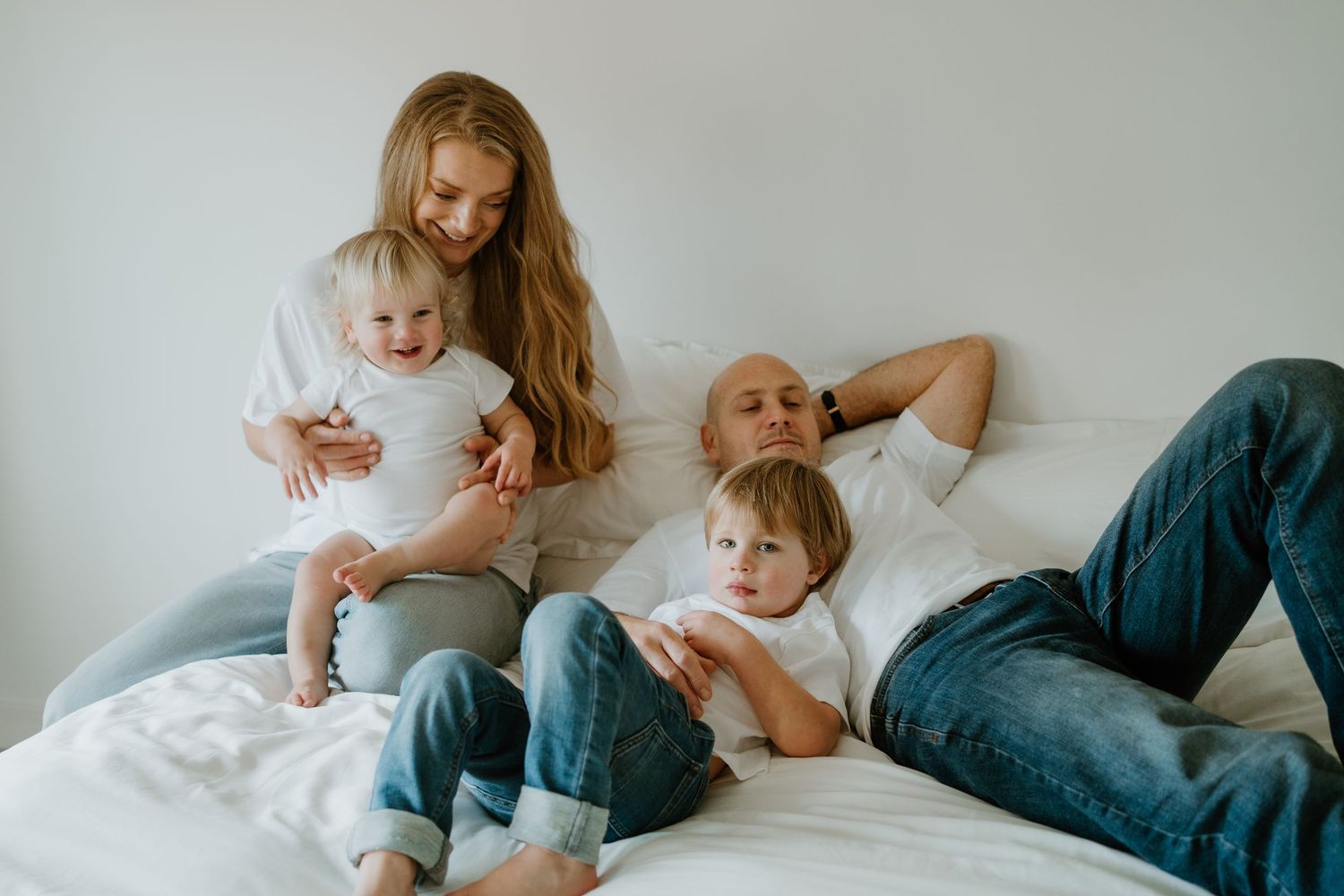 Young family relaxing together on white bed, mother holding baby while father and son lie down.