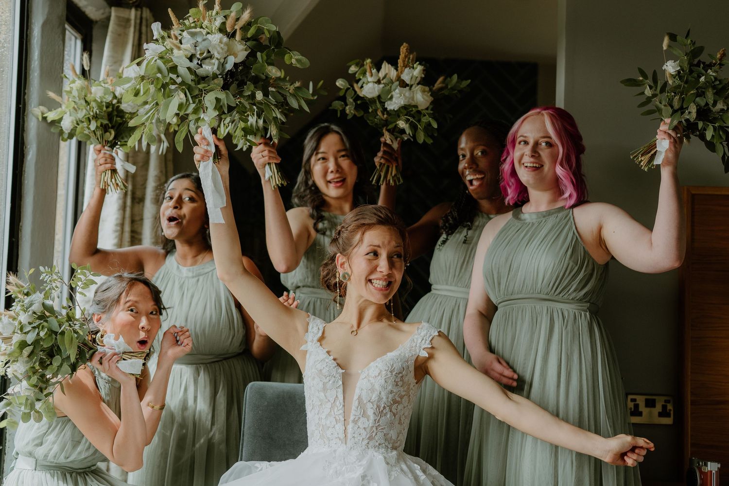 Bride and bridesmaids in sage green dresses celebrate joyfully raising bouquets.