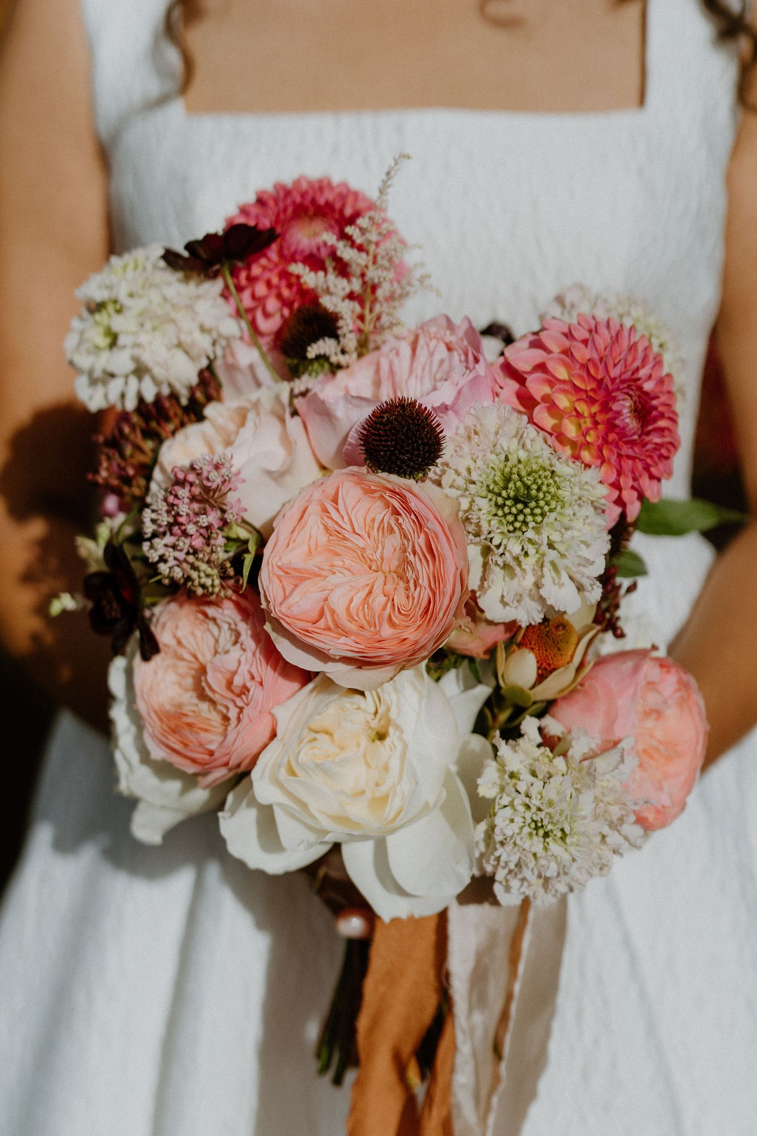 A bride holds a lush bouquet of pink dahlias, garden roses, and white blooms with ribbon streamers.