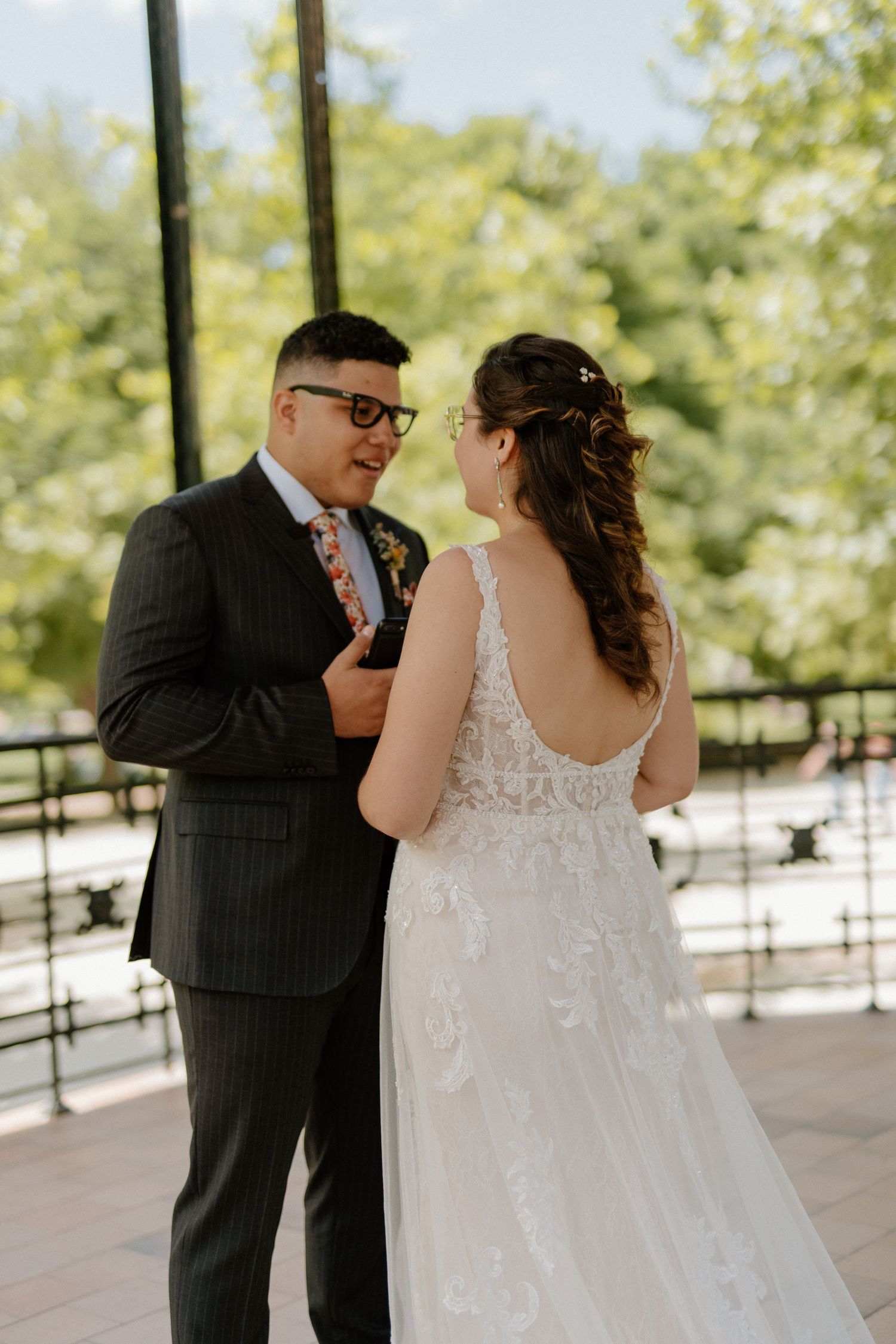 Groom in pinstripe suit smiling at bride in lace gown during their first look at outdoor venue.