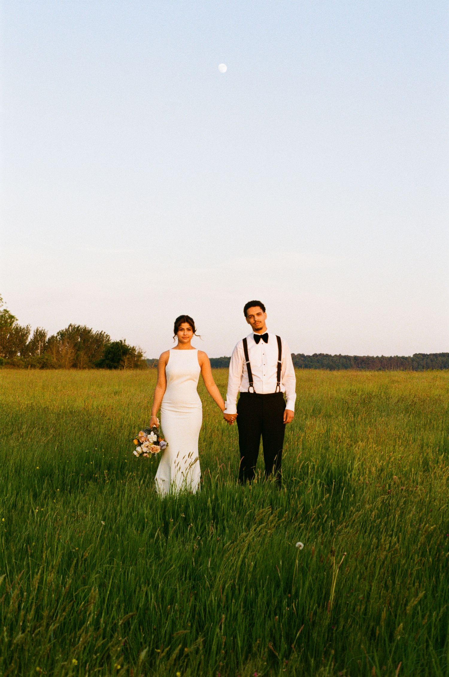 Bride and groom holding hands in lush green field at dusk with full moon above.