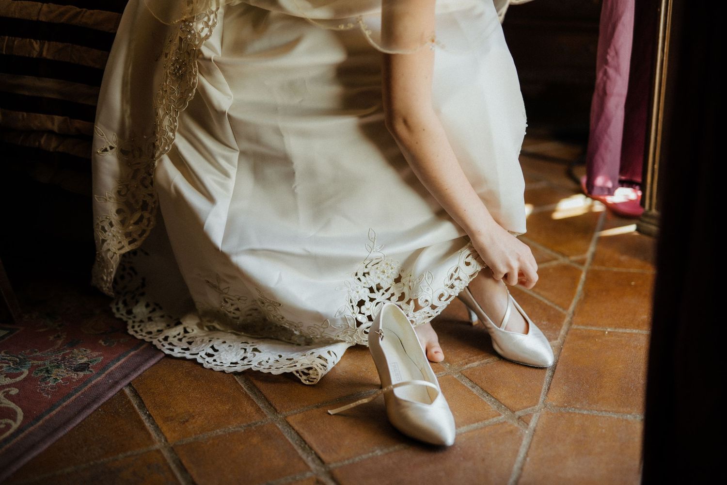 Bride in lace wedding dress putting on white satin heels on terracotta tile floor.