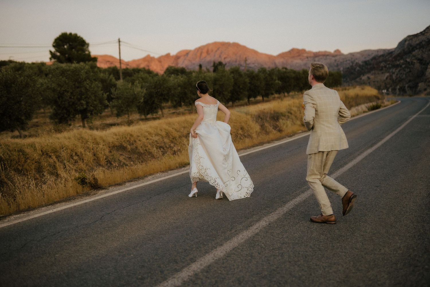 Bride and groom walking hand in hand down open road at sunset with mountain scenery.
