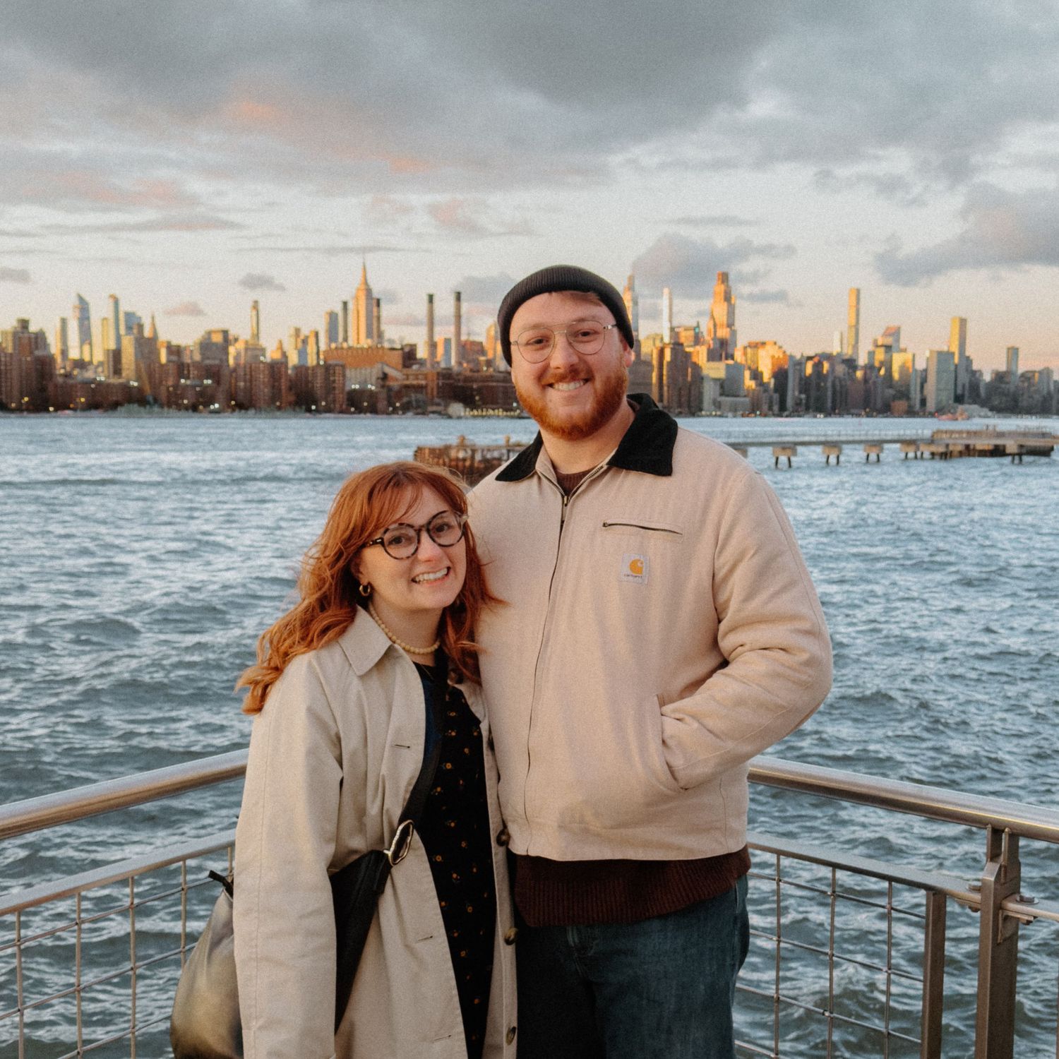 A smiling couple poses on a waterfront pier with the New York City skyline at golden hour behind them.