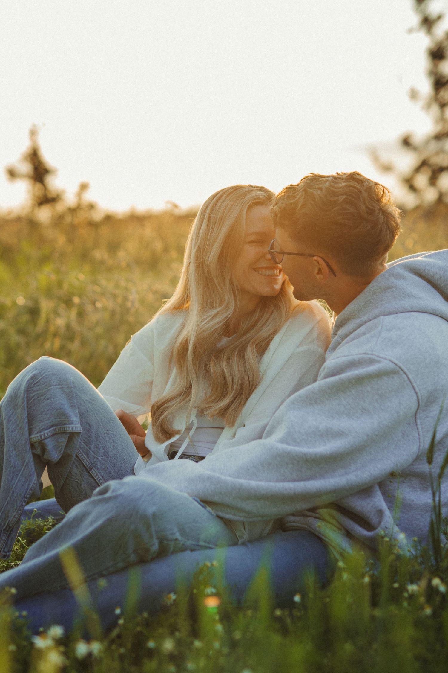 Romantic couple sits close together in golden sunset light in a lush green meadow field.