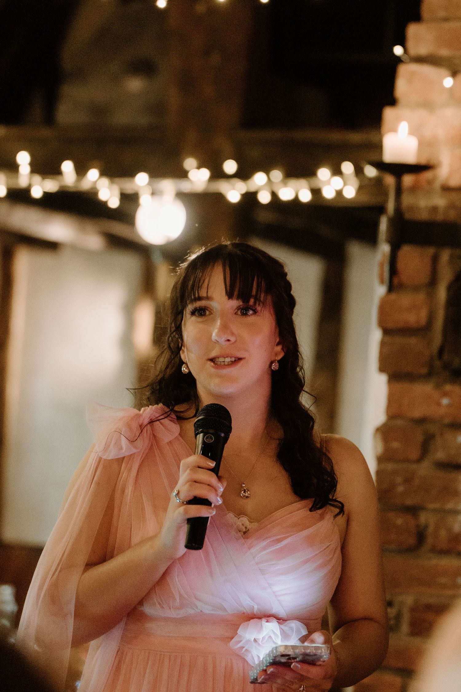 A bridesmaid in a pink tulle dress delivers a heartfelt speech with a microphone at a wedding reception.