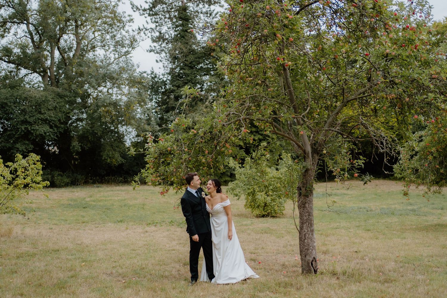 Bride and groom stand together in scenic meadow beside tree during outdoor wedding portraits.