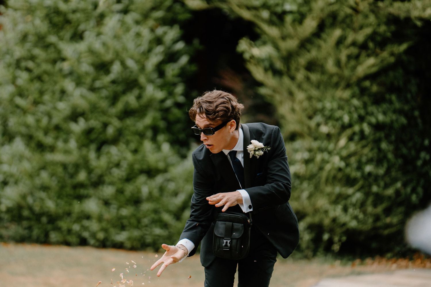 Young groomsman in dark suit with boutonniere tosses confetti on garden pathway at wedding ceremony.