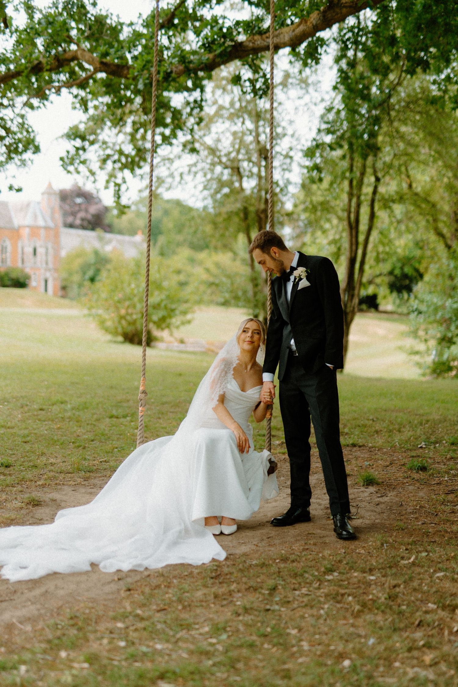 Bride sitting on a rope swing under a tree as groom stands beside her in a lush garden wedding setting.