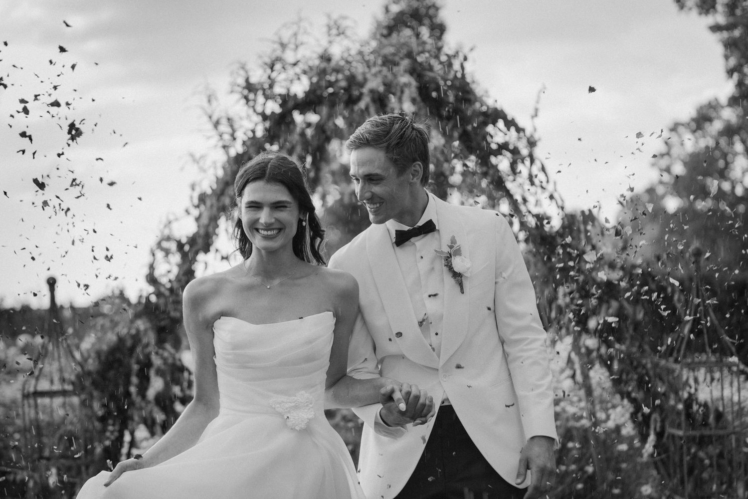 Black and white photo of bride and groom celebrating with confetti under outdoor floral arch.