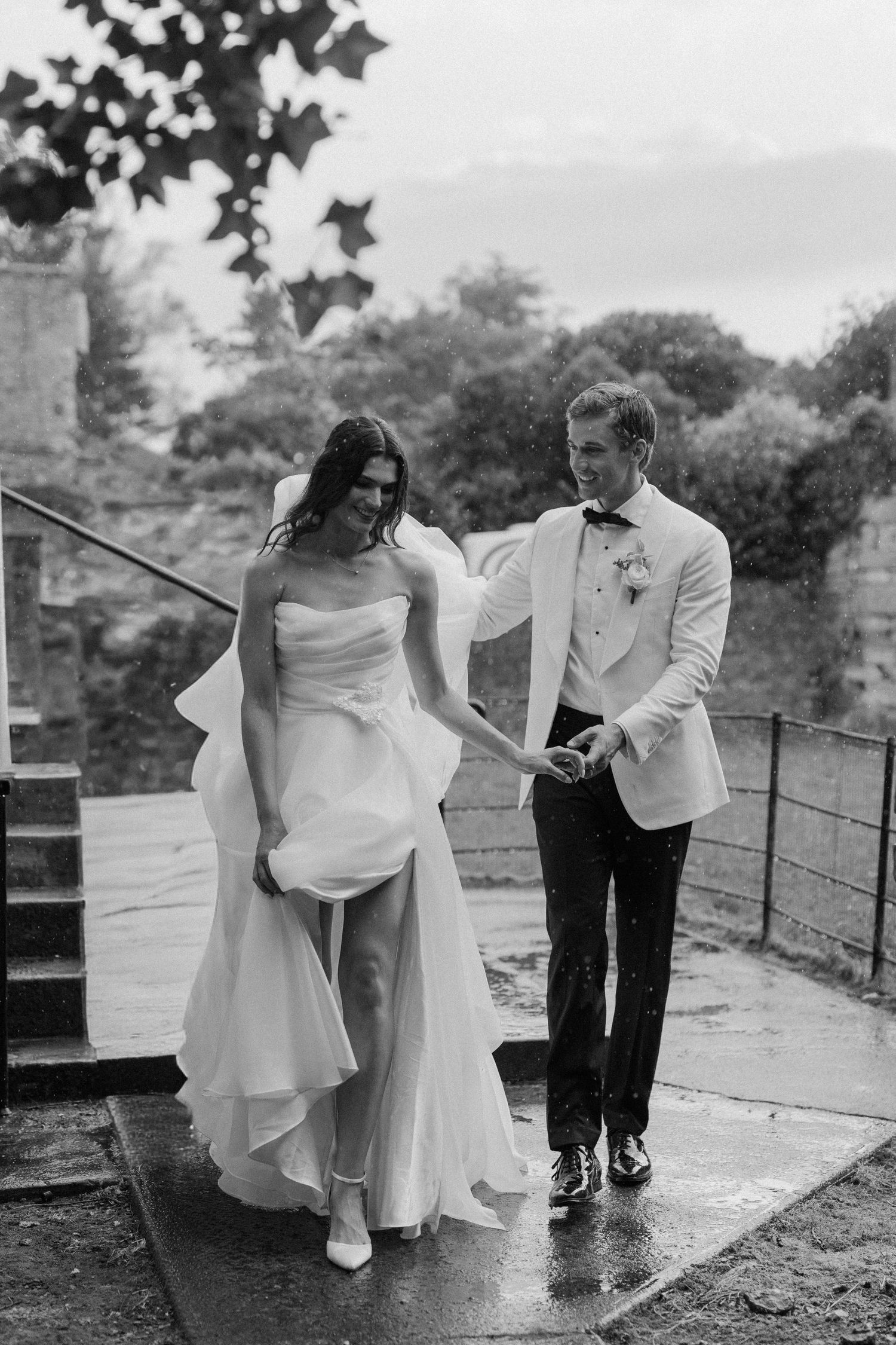 Black and white photo of bride and groom walking hand in hand in the rain outside a historic stone venue.