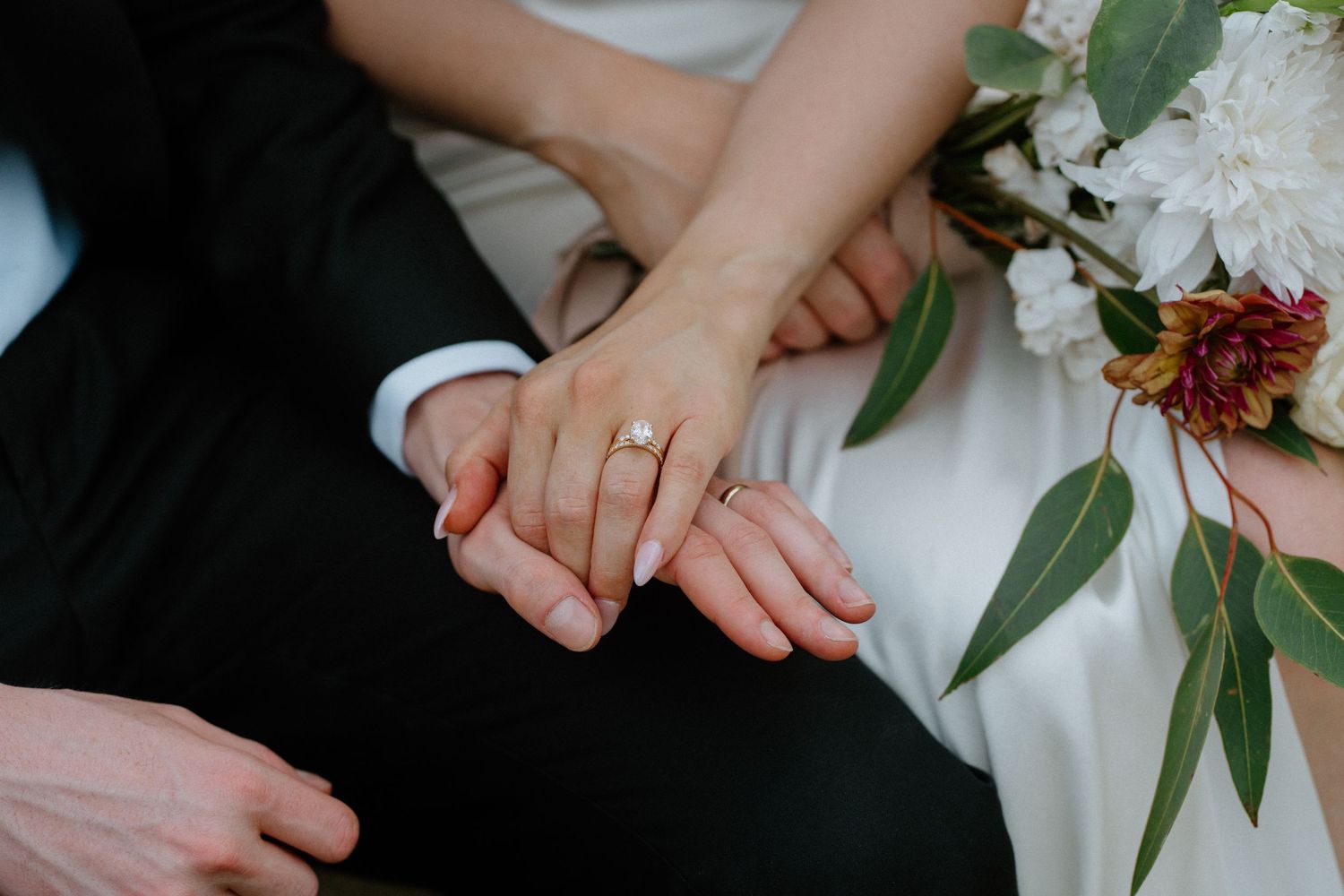 Close-up of newlyweds hands showing gold wedding ring with bridal bouquet in background.