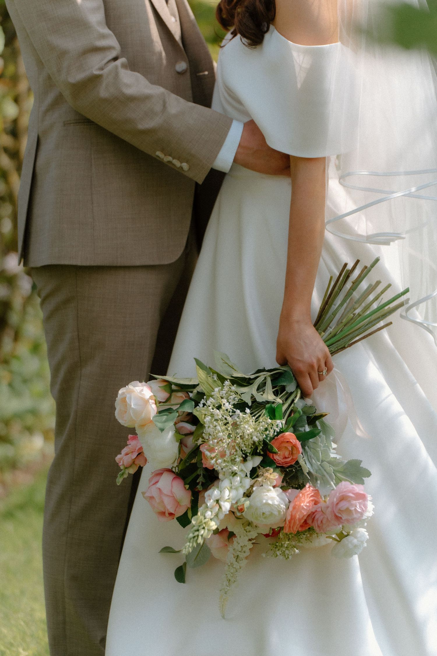 Close-up of bride and groom embracing, bride holding lush pink and white peony wedding bouquet.