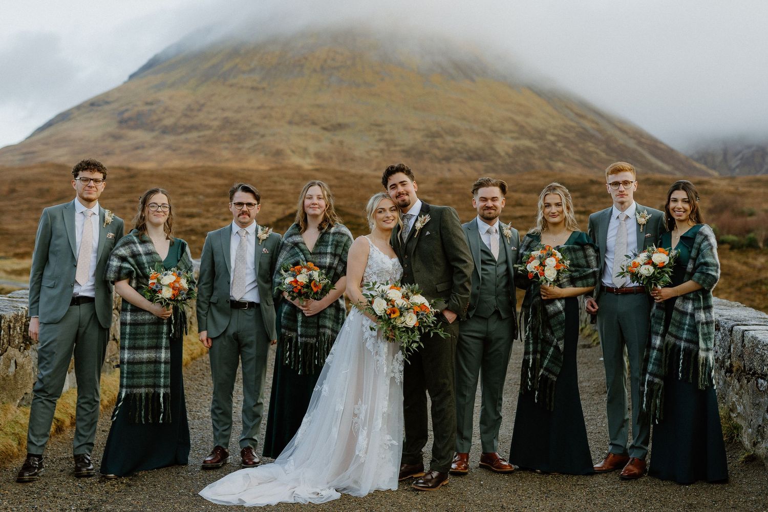 Wedding party in tartan shawls poses before dramatic misty Scottish highland mountain backdrop.