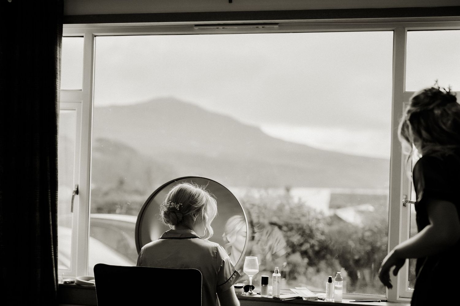 Black and white image of bride at vanity mirror with mountain view through large window.
