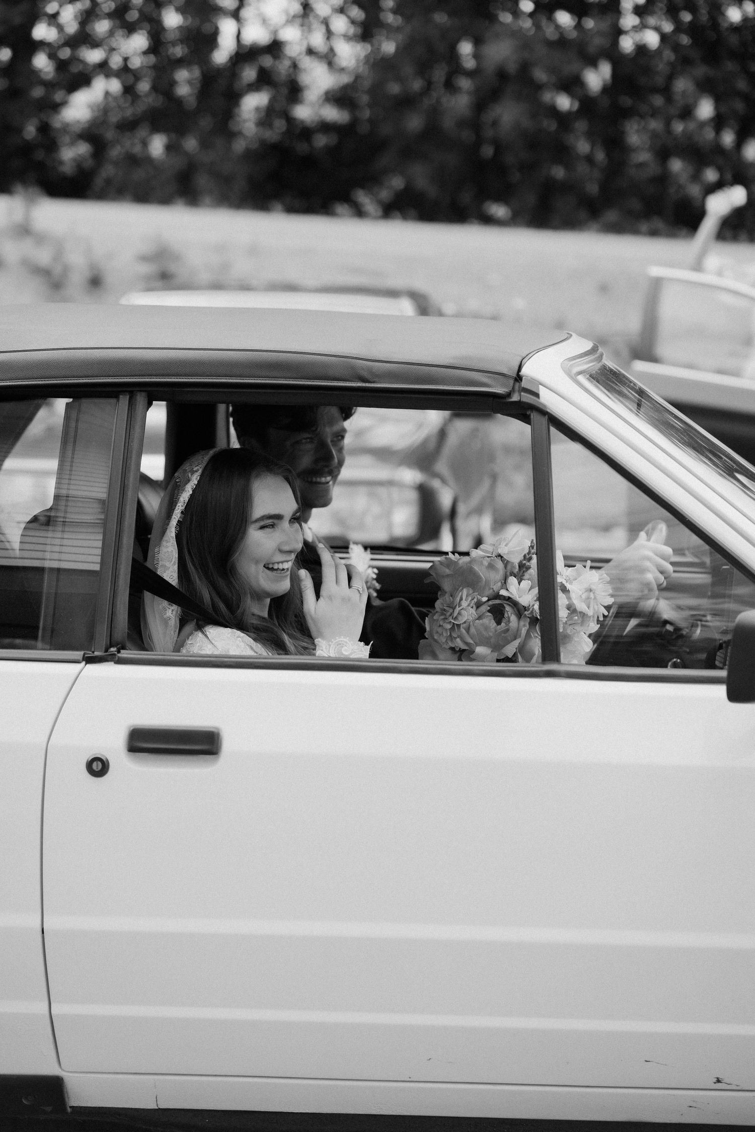Black and white photo of laughing bride in veil sitting in white convertible with groom.