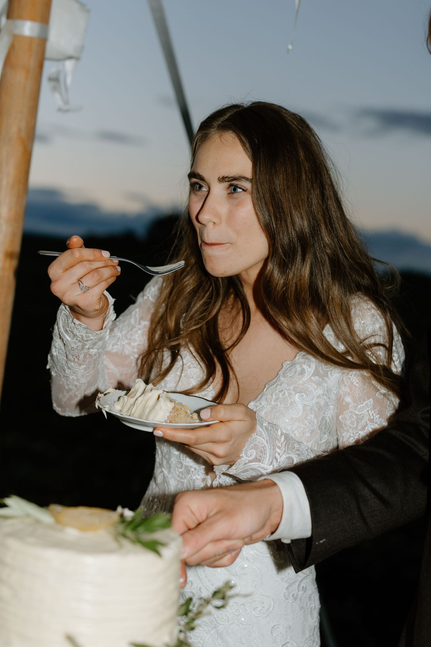 Bride in lace gown tasting wedding cake at outdoor twilight reception.