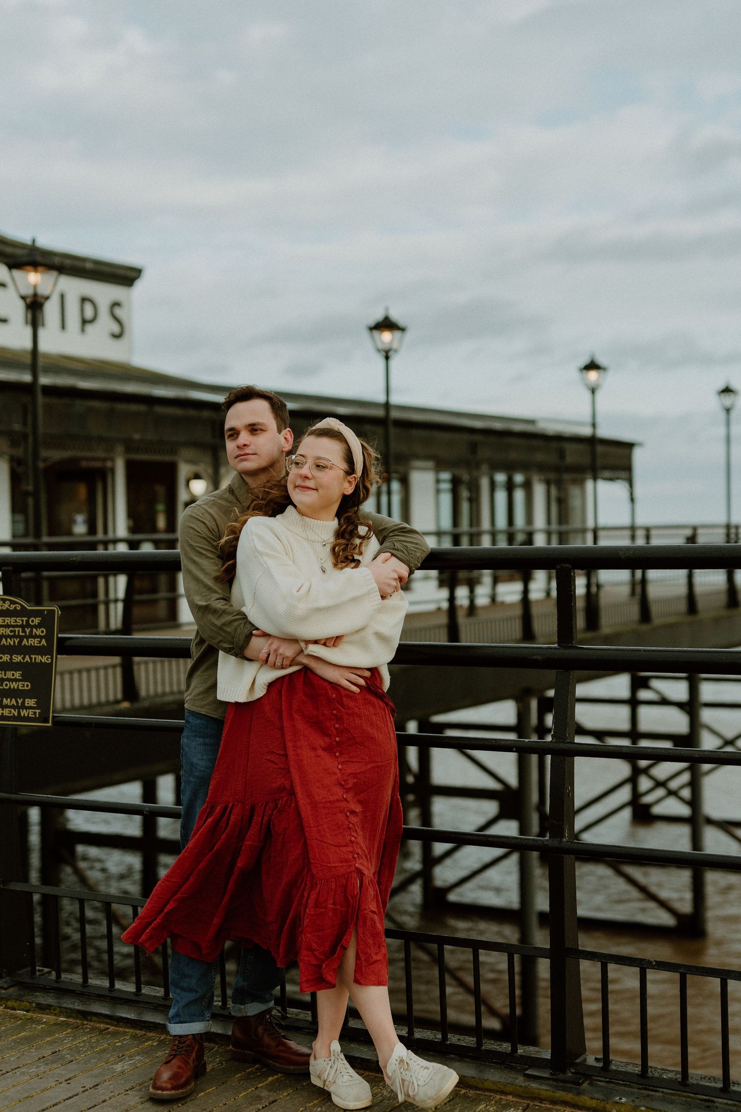 Couple embracing near a pier, woman in red skirt and white sweater with vintage lamps.