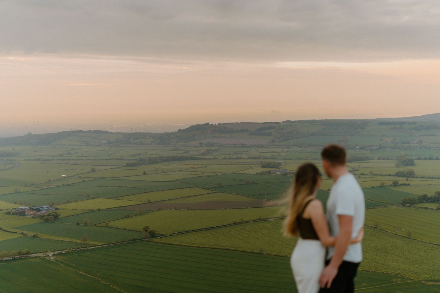 Couple standing together overlooking a vast green patchwork countryside at sunset.