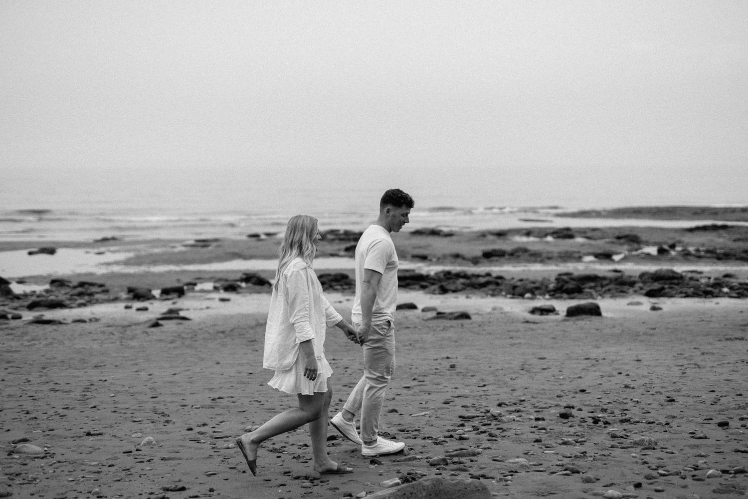 Black and white photo of a couple holding hands walking along a rocky beach shoreline.
