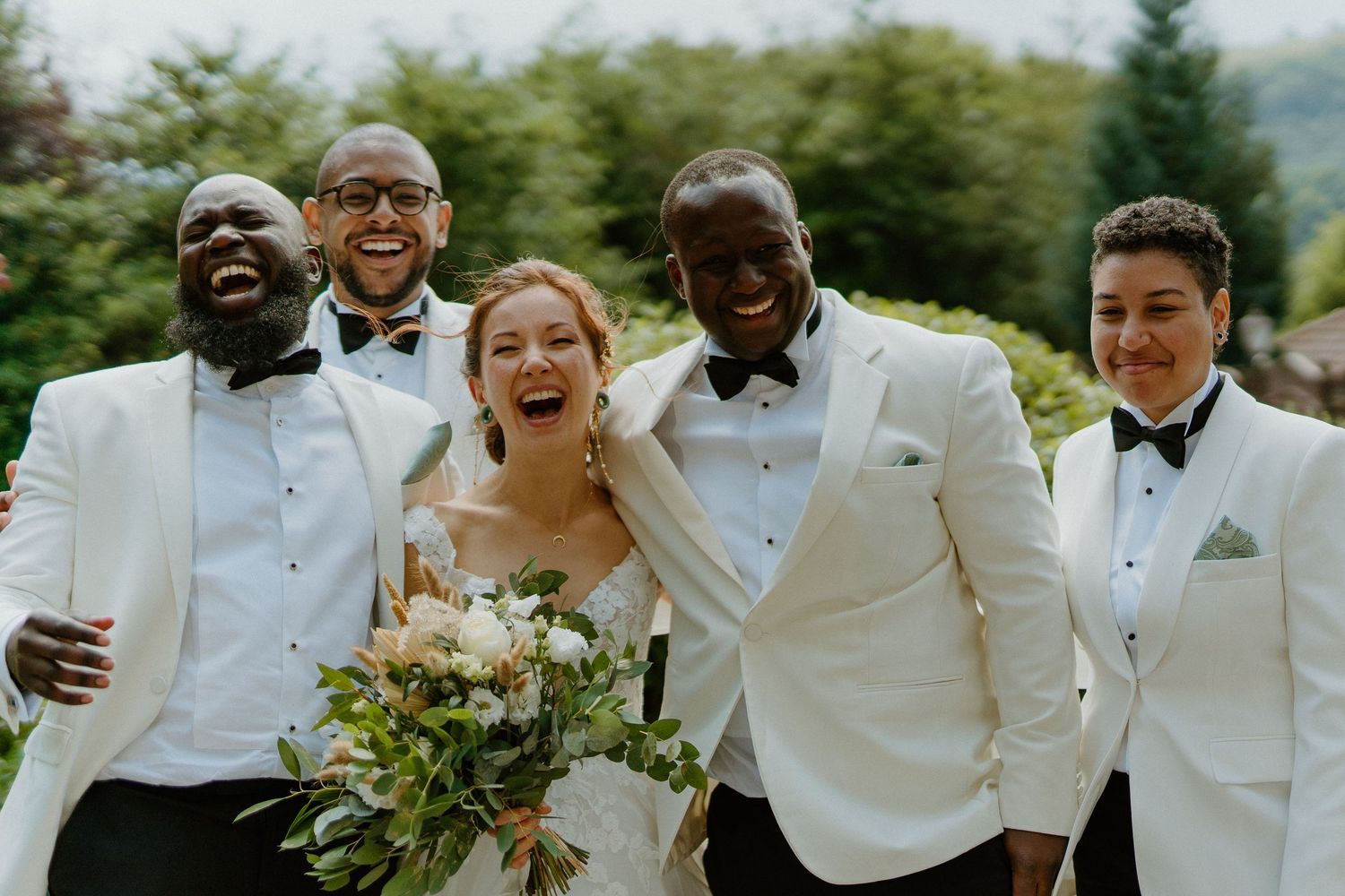 Bride laughing with four groomsmen in white tuxedos during outdoor wedding portrait.