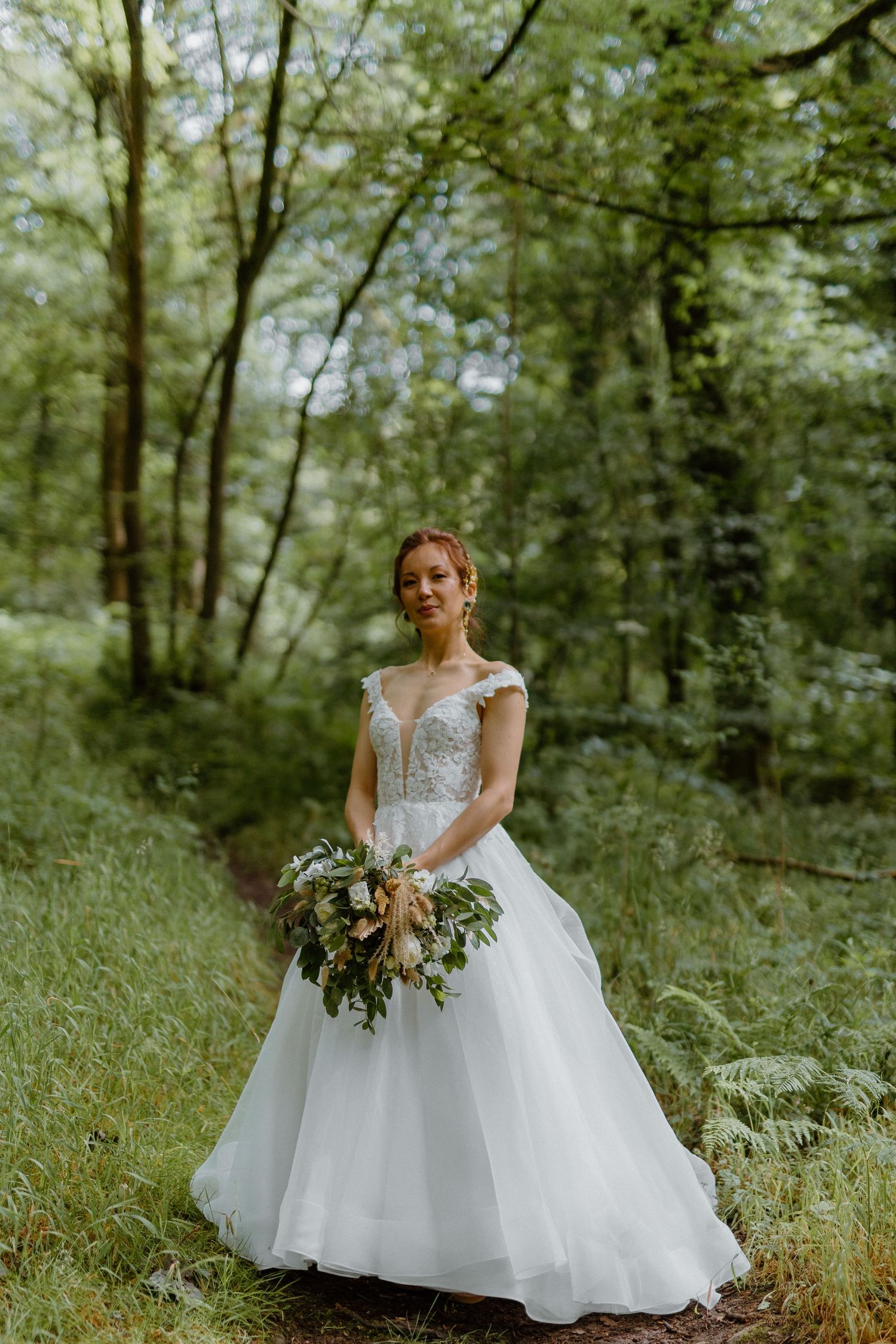 Bride in white lace ballgown holding bouquet of greenery while standing in lush forest setting.