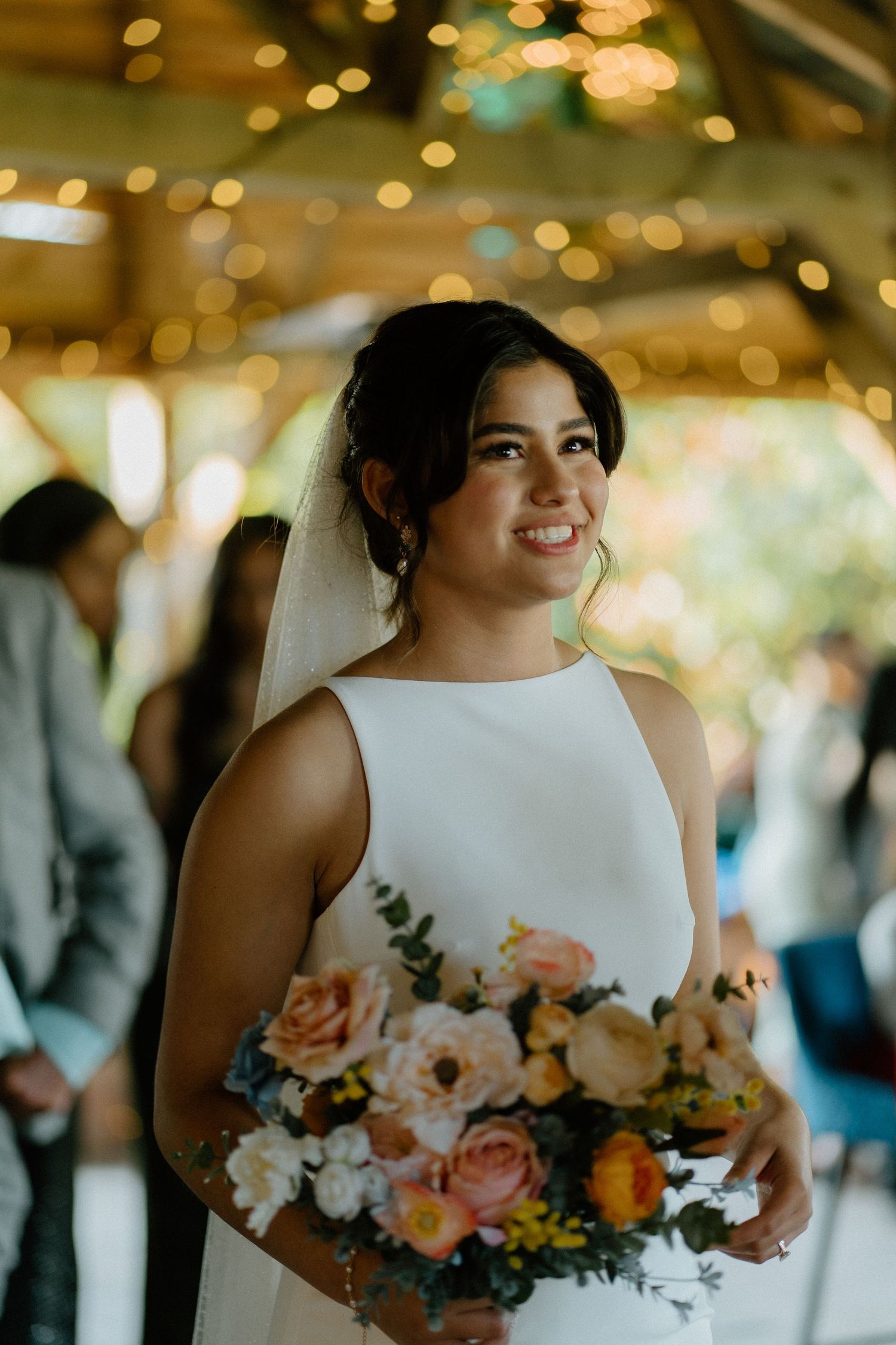 Beautiful bride in white halter gown smiles while holding lush peach and pink floral bouquet with bokeh lights.