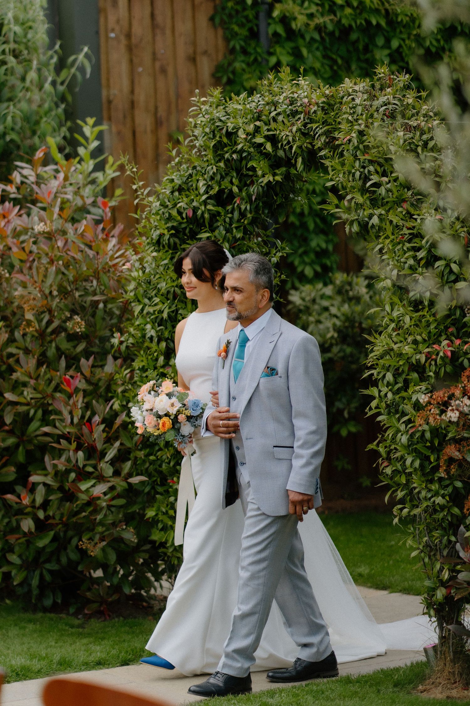 Bride in elegant white gown walking down the aisle with her father through a lush garden setting.