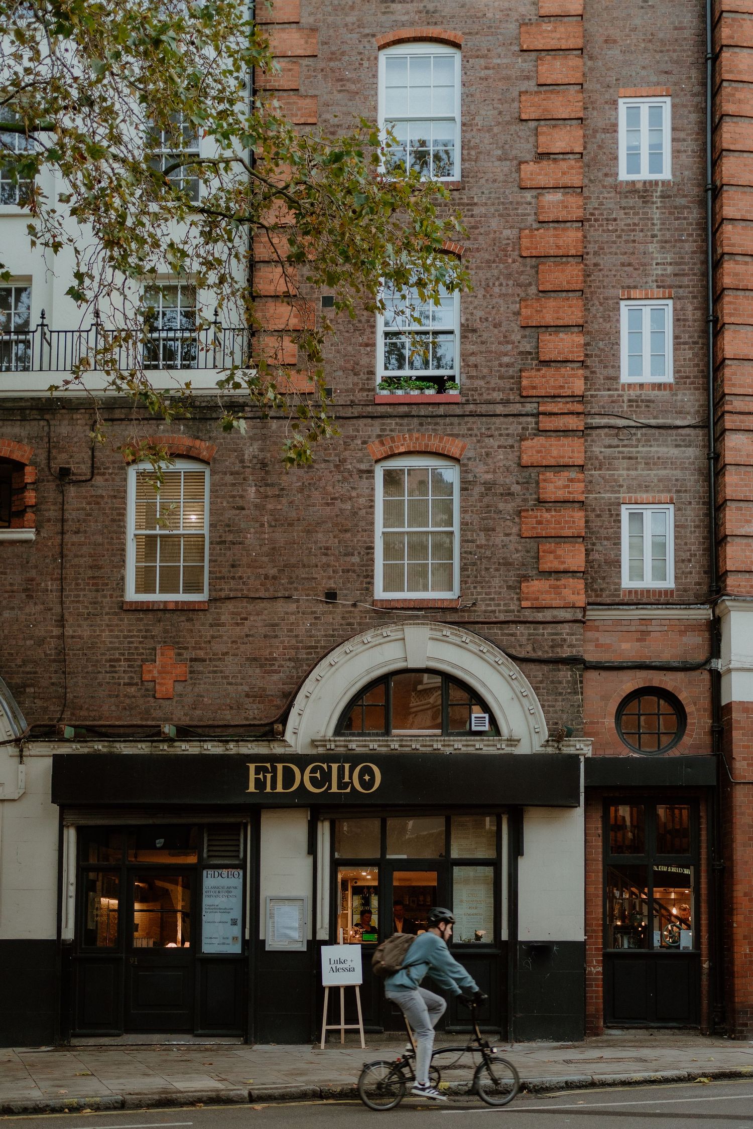 Exterior of Fidelo restaurant in a red brick building with a cyclist passing on the street.
