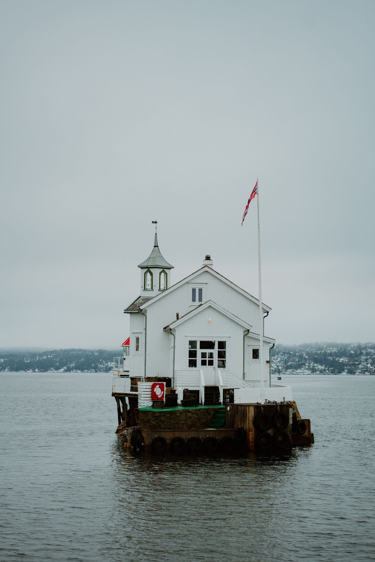 A small white lighthouse chapel sits on a wooden platform in the middle of calm waters under an overcast sky.