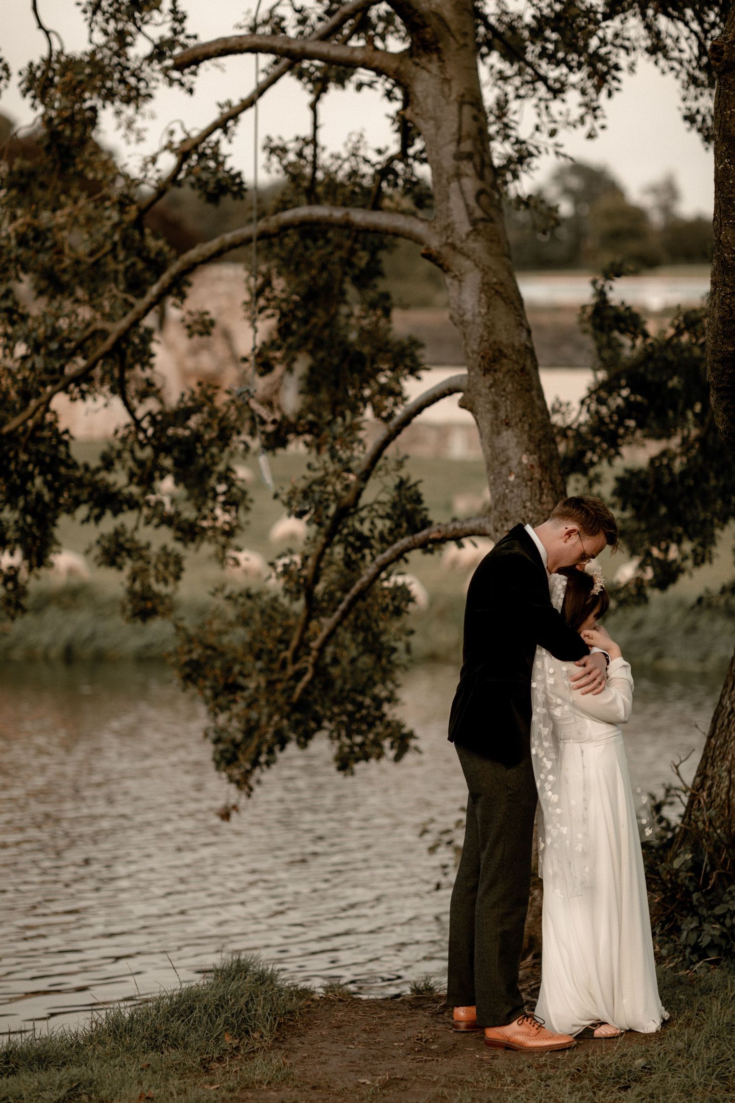 A romantic wedding couple embraces beneath a tree by a serene lake at sunset.