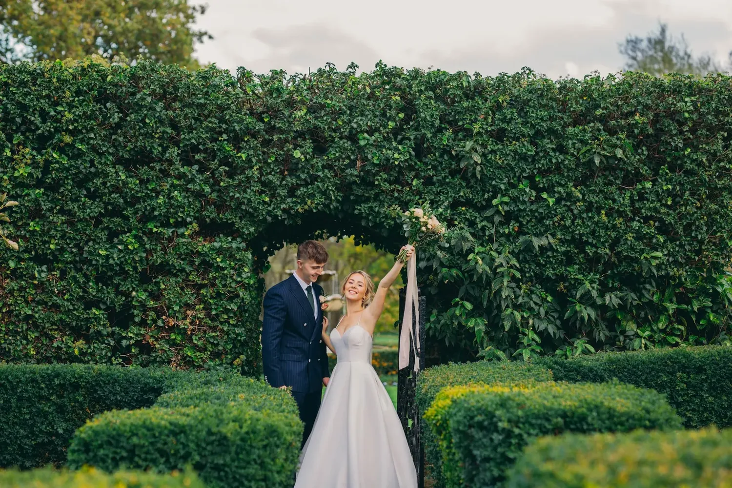 A bride and groom celebrate in a manicured garden with tall green hedges.
