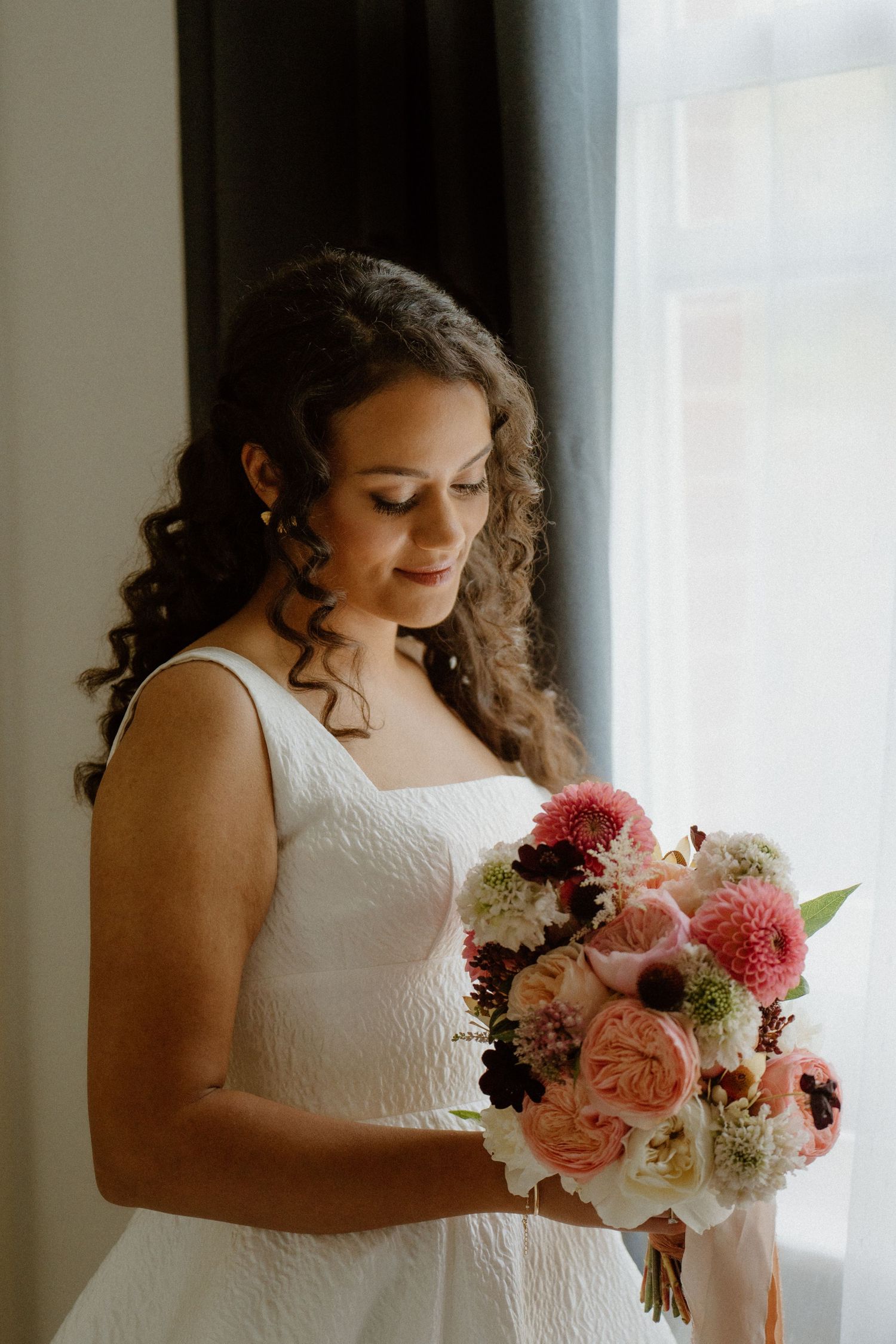 Beautiful bride with curly hair holding a stunning pink and white floral bouquet near a bright window.