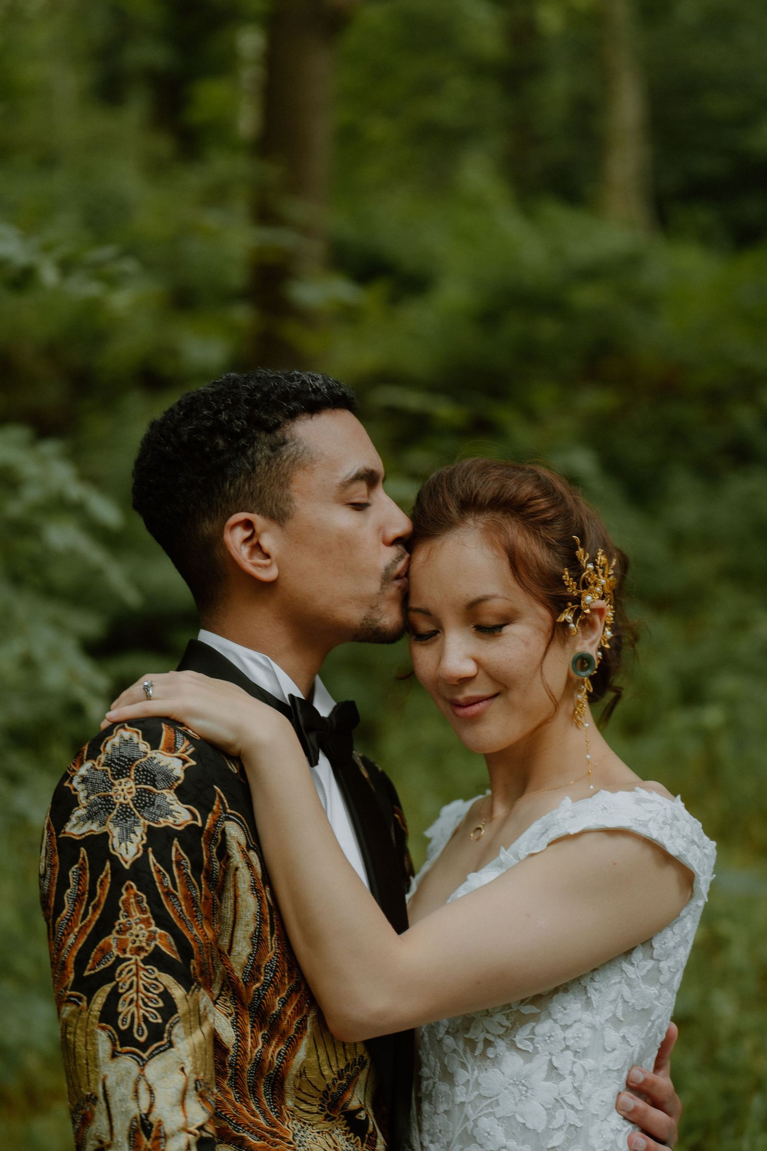Groom in gold batik jacket kisses bride on forehead in a lush green forest setting.