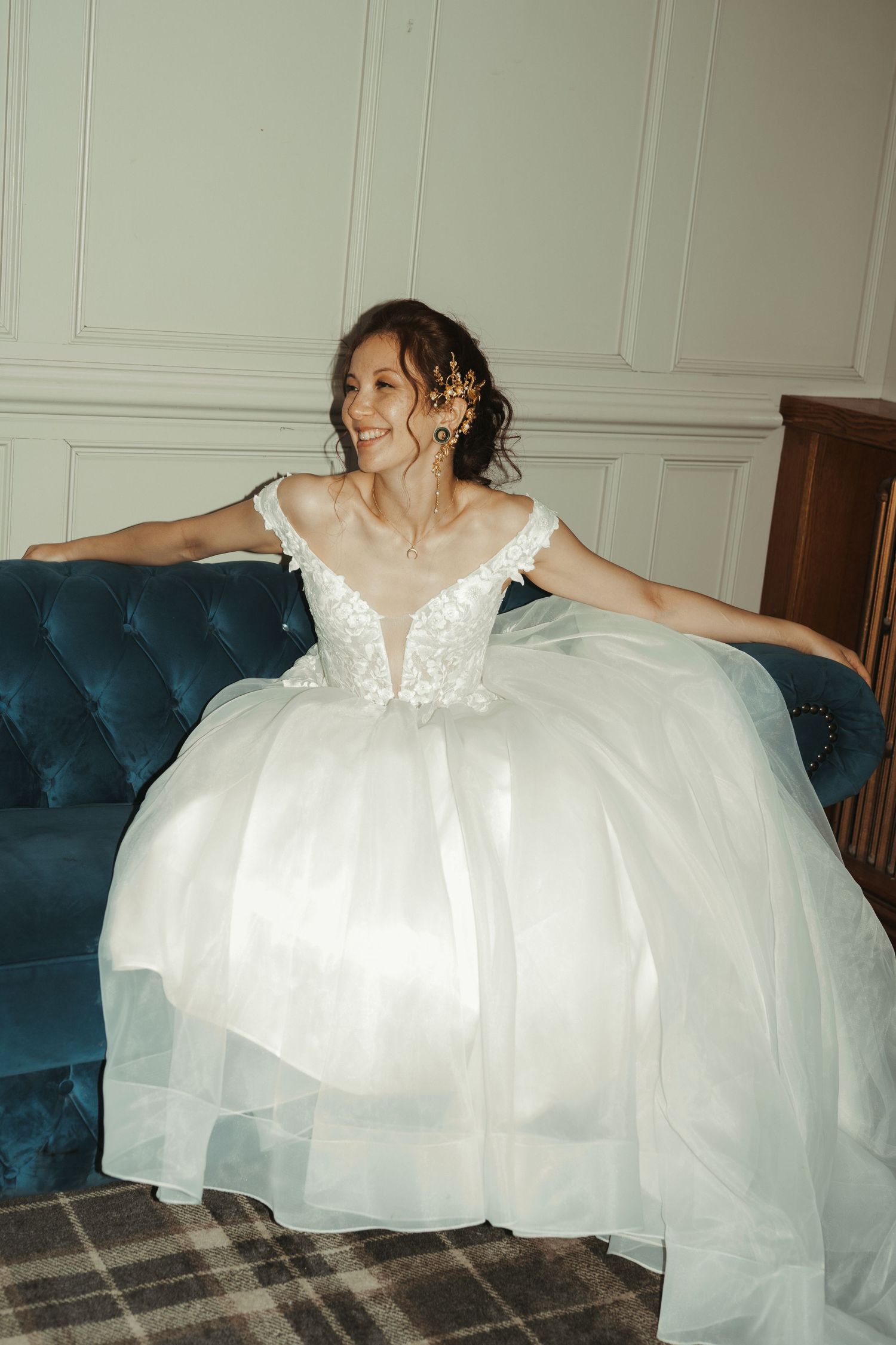 Smiling bride in off-shoulder ballgown with floral hair accessory seated on blue velvet sofa.