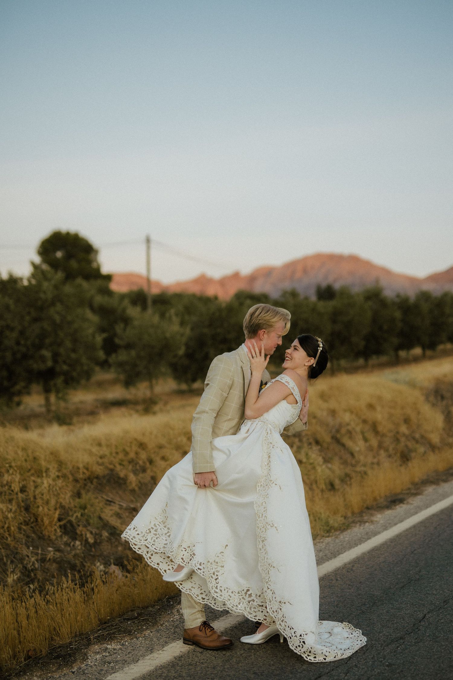Groom dipping bride in wedding dress roadside at golden sunset with scenic mountain views.