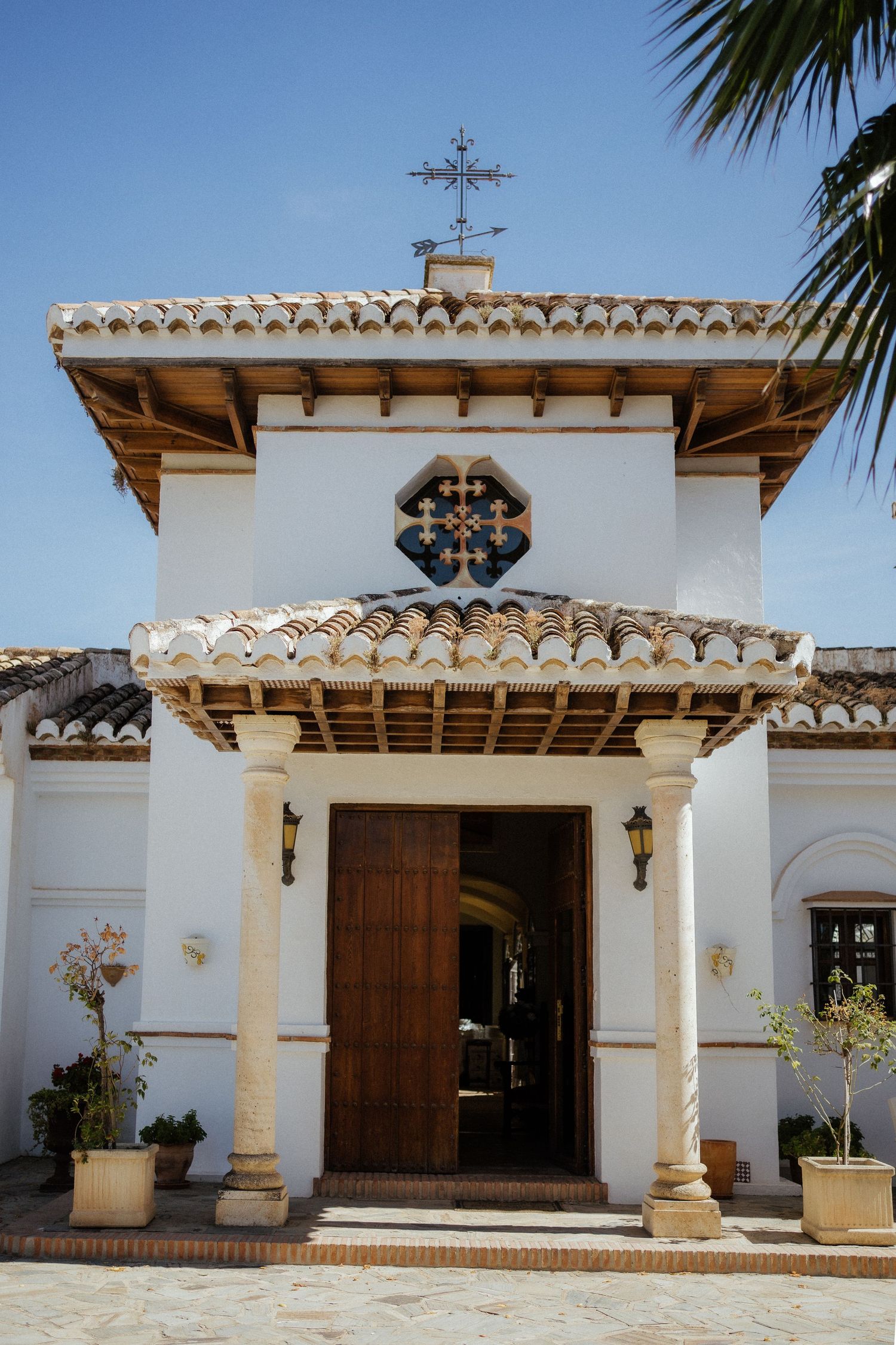 White Spanish colonial chapel entrance with wooden door, terracotta roof tiles, and decorative cross under blue sky.