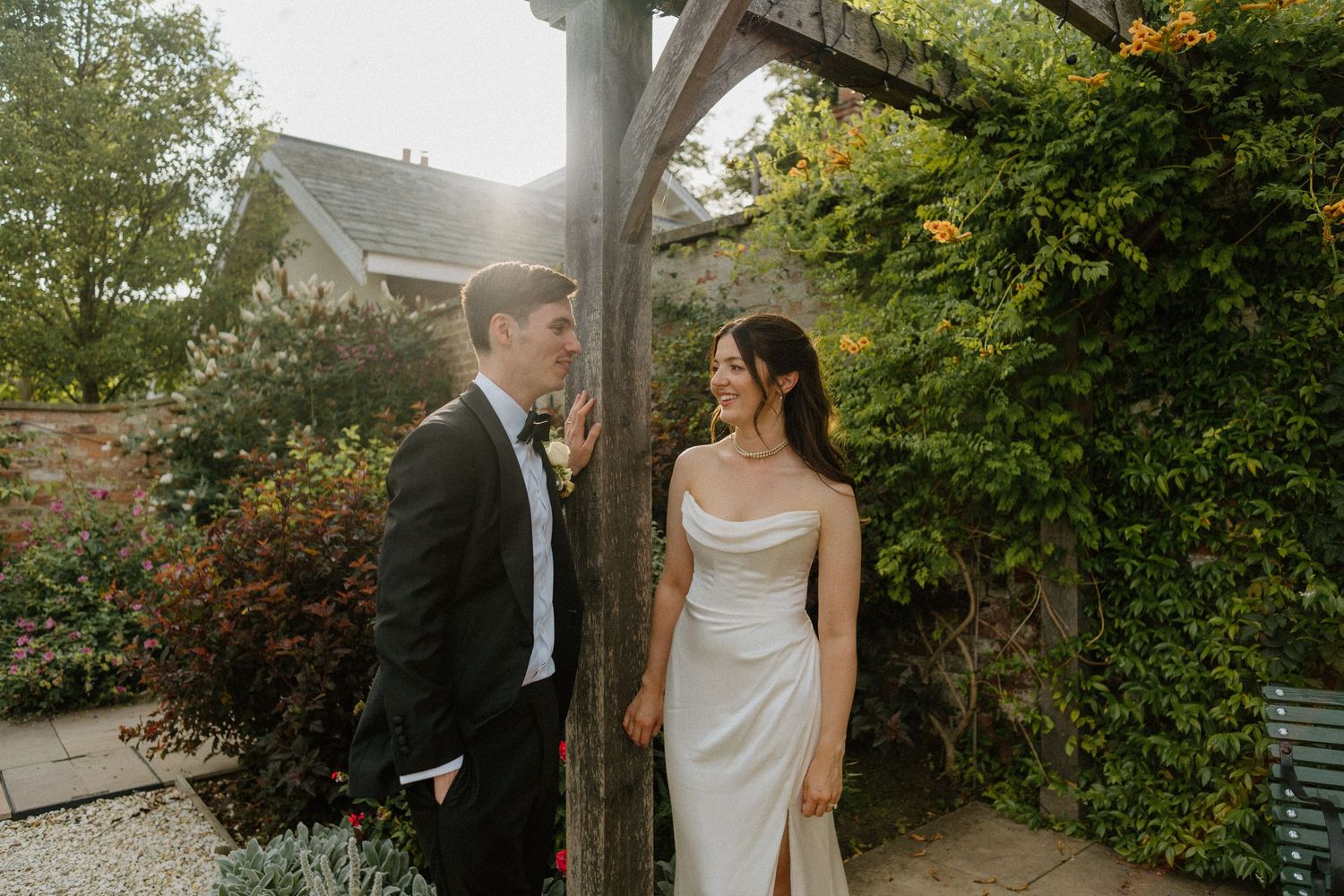 Bride in a cowl-neck gown smiling at her groom in a sunlit garden pergola setting.