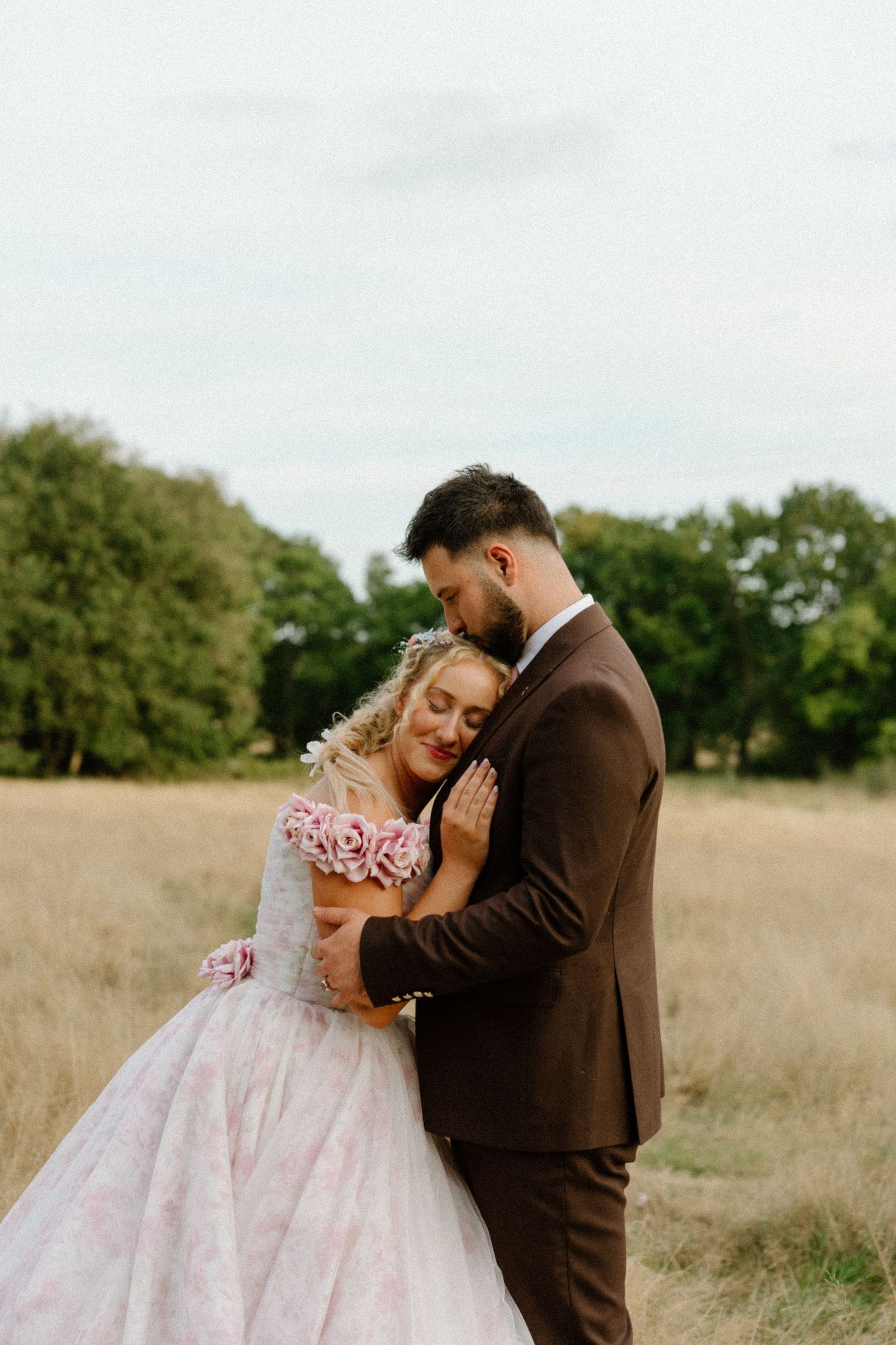 A bride in a floral pink gown embraces her groom in a golden meadow during couples portraits.