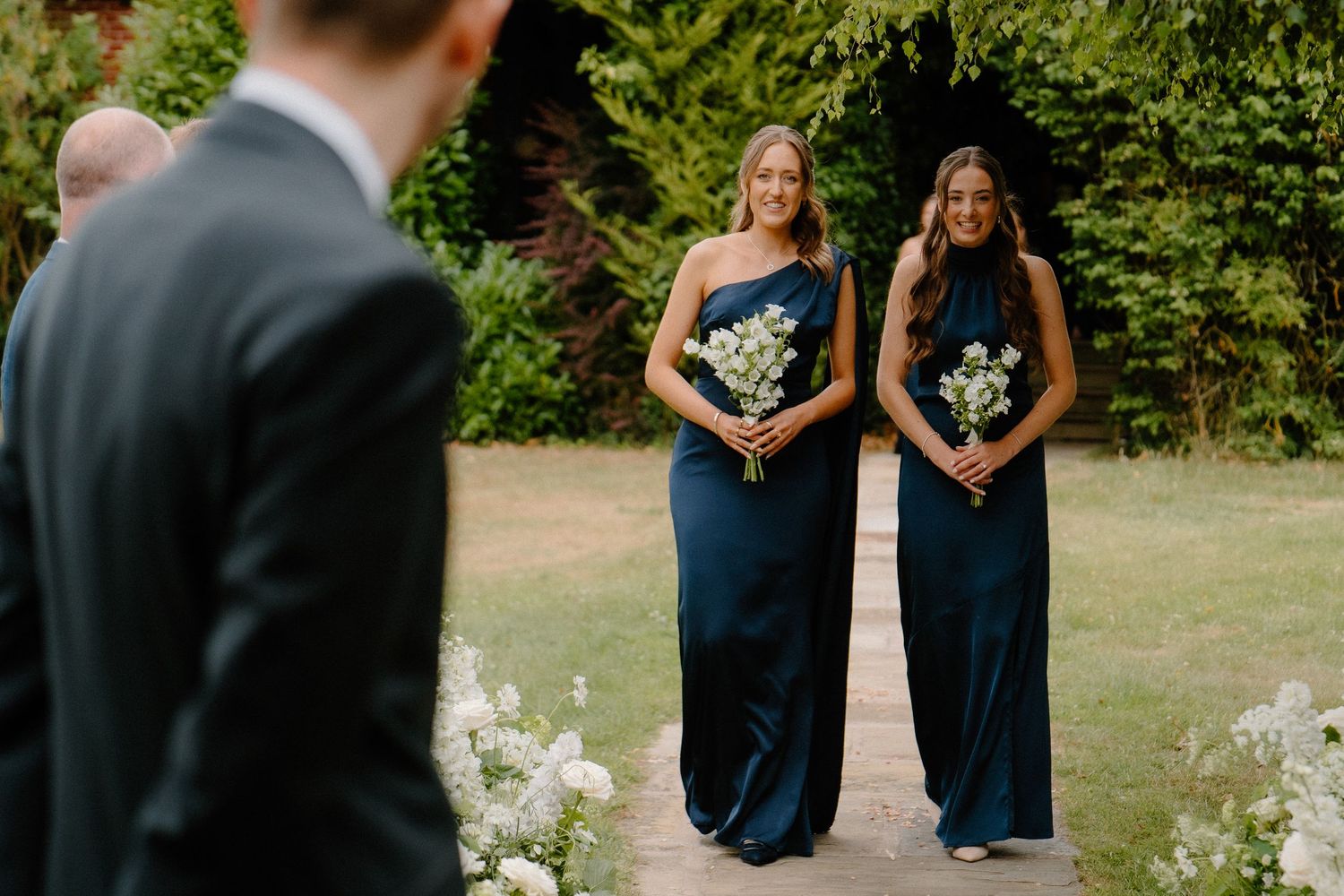 Two smiling bridesmaids in navy satin gowns hold white bouquets as they walk down the wedding aisle.