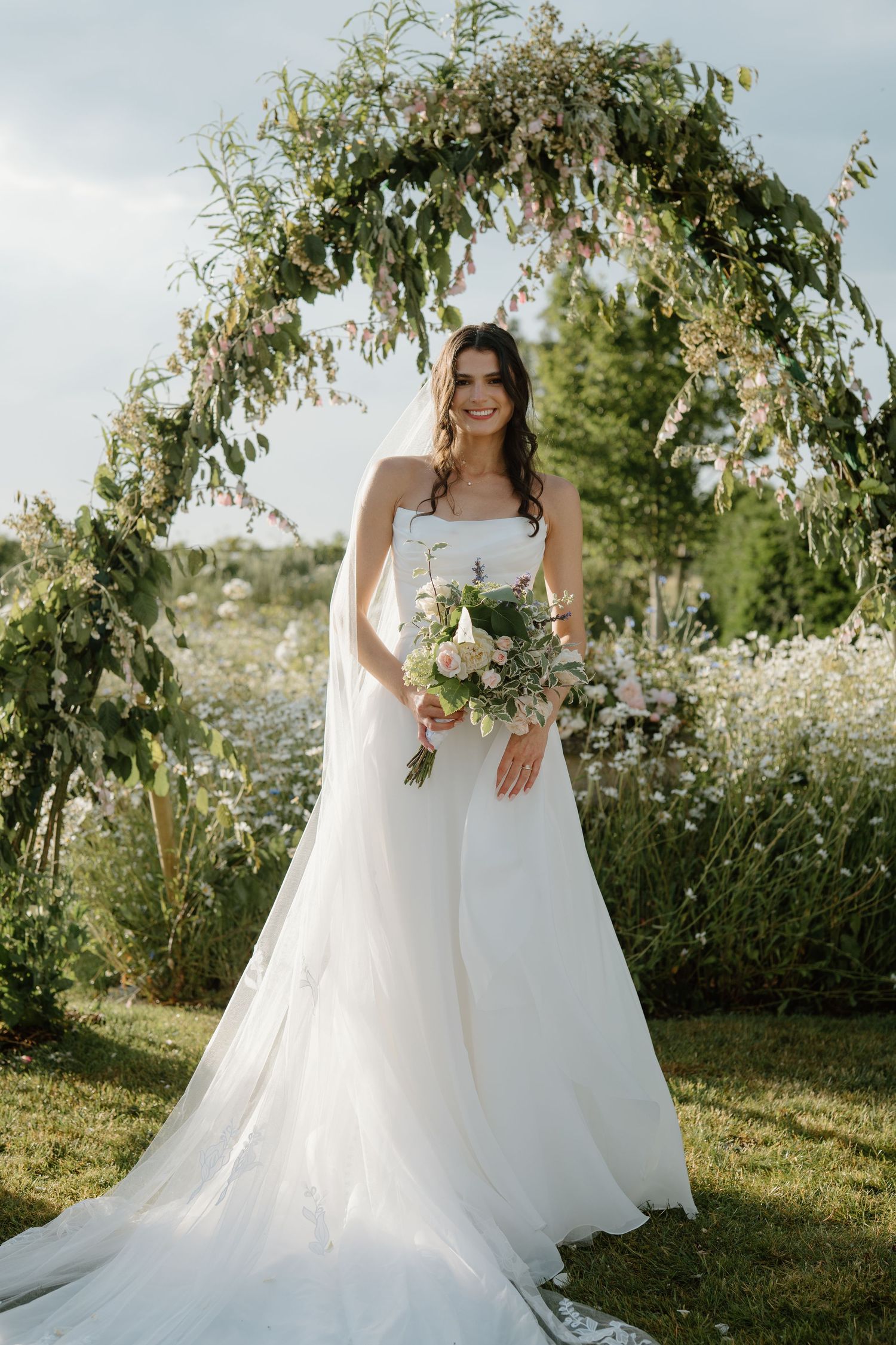 Radiant bride in strapless white gown holding bouquet under lush floral arch in garden.