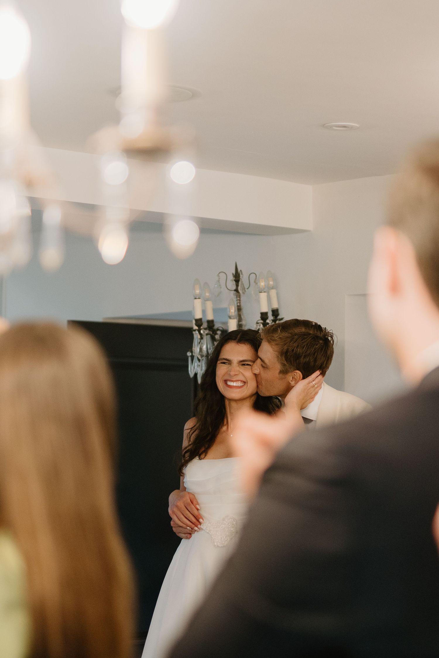 Groom kissing bride on cheek as she smiles during indoor wedding reception with chandelier.