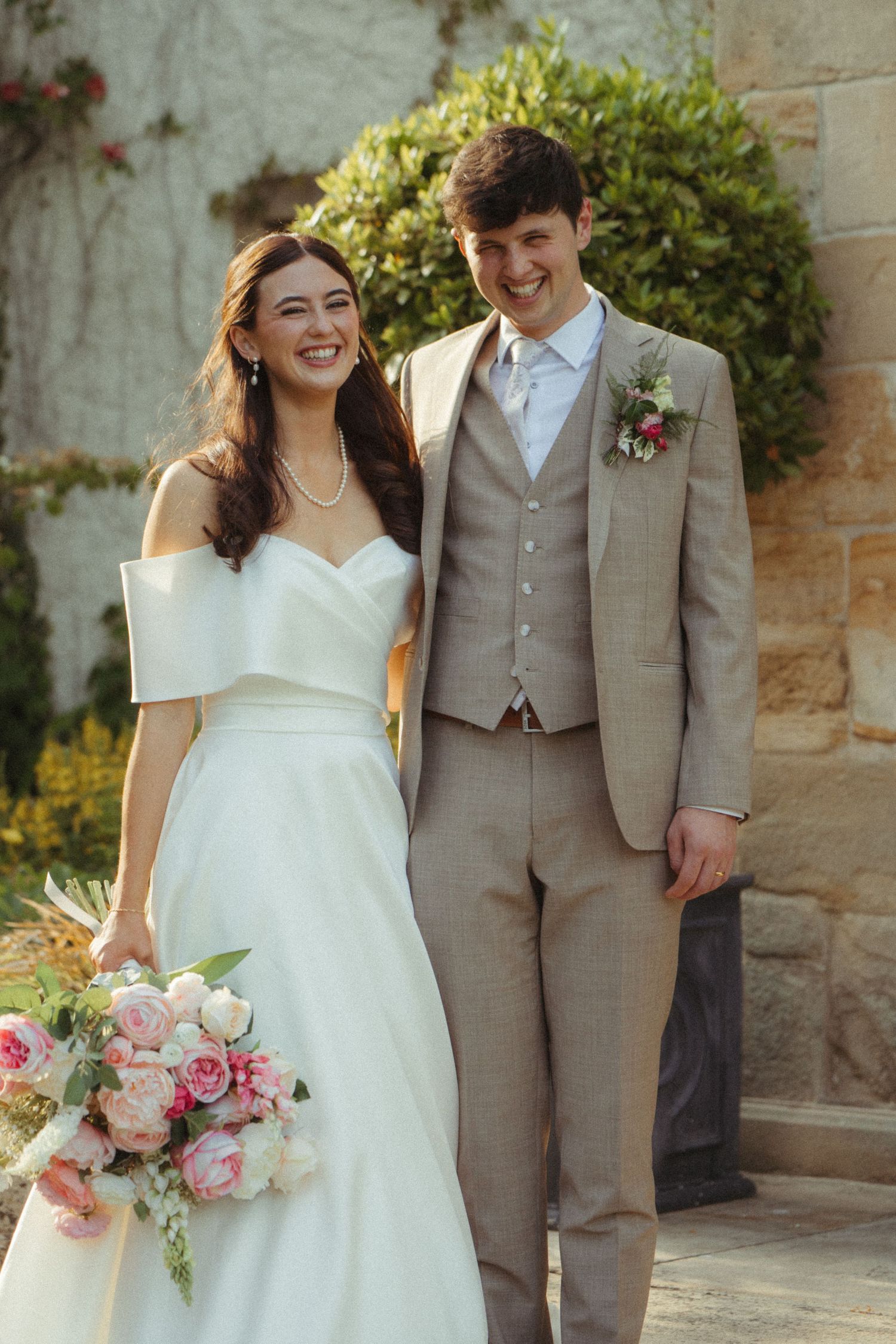Smiling bride in off-shoulder white gown and groom in grey suit walking outside stone building.