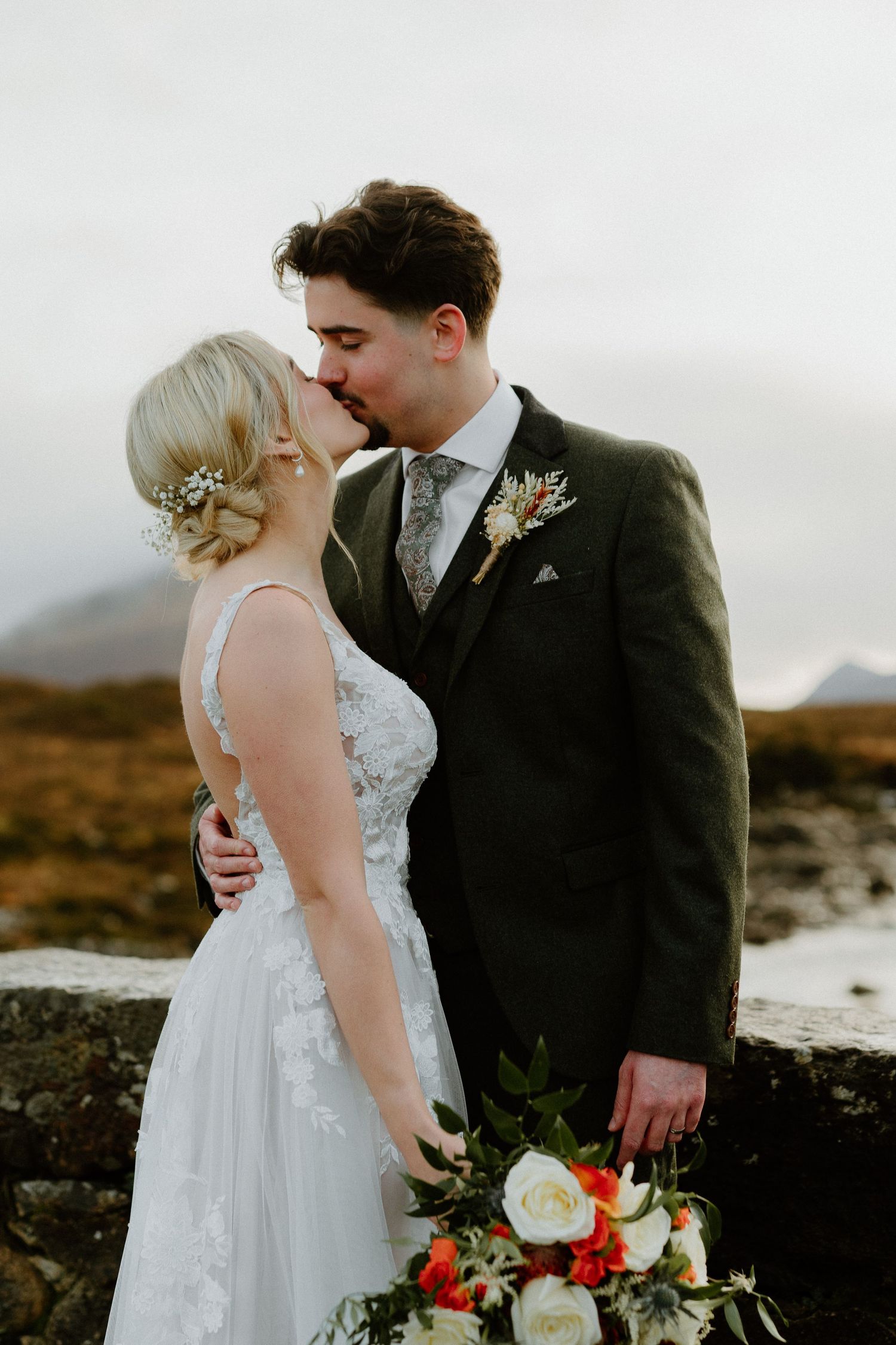 Bride and groom share romantic kiss in Scottish Highlands, bride holding vibrant orange and white bouquet against moorland backdrop.